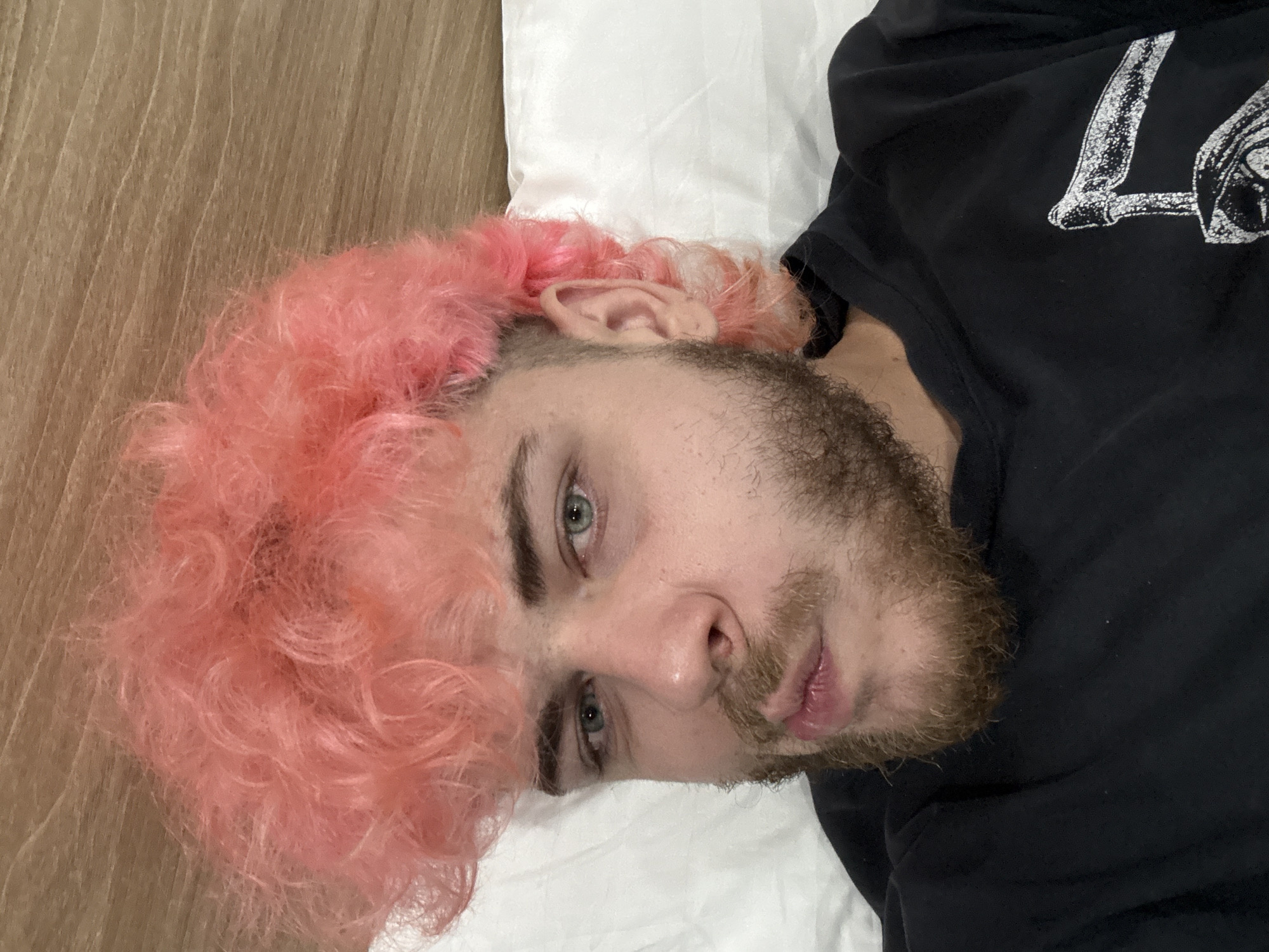 The image features a young man with light skin, light blue or green eyes, and a light brown beard and mustache. He is lying down, positioned diagonally in the frame, and looking directly into the camera with a neutral expression. His most prominent feature is his bright, curly, bubblegum pink hair.
He is wearing a black t-shirt with a white graphic print on the chest. A partial view of this graphic is visible, appearing as an angular, distressed letter or symbol, possibly an 'L' or part of a larger design, but not fully legible as text. The man's head rests on a plain white pillow, which occupies the lower right portion of the image. To the left and upper left, a light brown wooden surface with a distinct vertical grain pattern is visible, suggesting a floor, wall, or headboard in an indoor setting. The lighting appears to be ambient, likely indoor. No specific visual cues within the image indicate the provided location context of Đà Nẵng, Vietnam.
