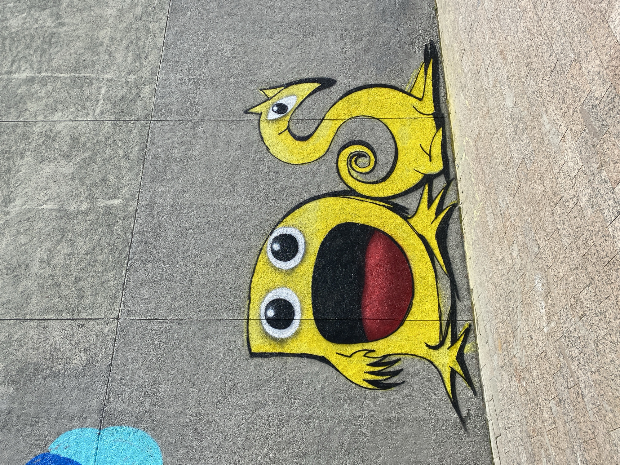 The image shows a yellow graffiti art piece on a gray concrete wall. The graffiti depicts a whimsical, cartoon-like creature. It has a large, round head with wide, white-rimmed eyes and a gaping mouth that is black with a touch of red inside. Above the head is a stylized yellow "S" shape with a smaller, bird-like head. The creature has pointy, black-outlined appendages extending outward. The artwork appears to be painted on an outdoor concrete wall, part of which adjoins a wall made of smaller, textured stones. In the lower-left corner, there is a partial view of blue graffiti. This art is located in Yekaterinburg, Russia.