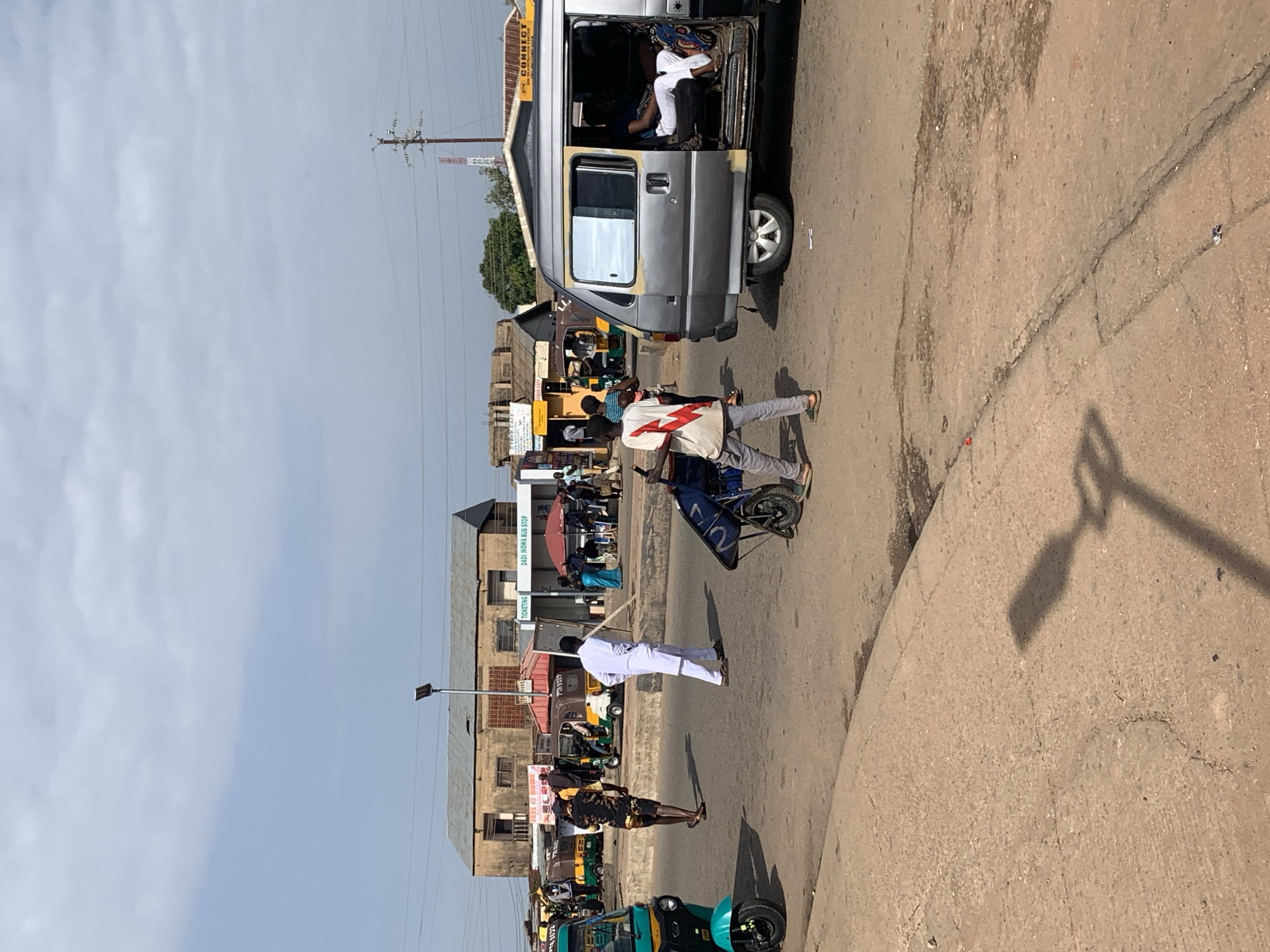 The image depicts a street scene in Gura Top, Nigeria, characterized by a dusty road and a bright, overcast sky. In the foreground, a person in gray pants and a white and red shirt pushes a blue cart. To the right, a silver minivan is parked with its side door open, revealing a person lying down inside. A pedestrian in white clothing walks away from the minivan, and another person in a black and yellow patterned shirt walks away from the camera towards the left. Several colorful auto-rickshaws are visible on the road, some occupied by passengers. In the background, a long, weathered building with a corrugated metal roof houses various businesses, including "CYBER CAFE" and "DADI IKOWA BUS STOP." A tall transmission tower stands in the distance. The overall impression is that of a busy, informal transportation hub in a tropical or subtropical urban environment.