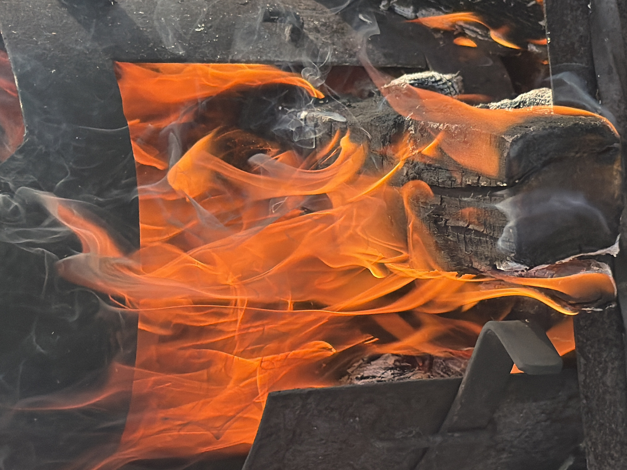 The image depicts a close-up view of a fire burning within what appears to be a metal brazier or fire pit. Intense orange and yellow flames dominate the frame, swirling upwards and casting a warm glow on the charred wood pieces within. Wisps of white smoke are also visible, mingling with the flames and rising towards the top of the image. The background is dark and somewhat out of focus, with hints of metal structures on the sides. The scene is devoid of any people or identifiable landmarks within Chaikovsky, Russia. The focus is entirely on the raw power and movement of the fire.
