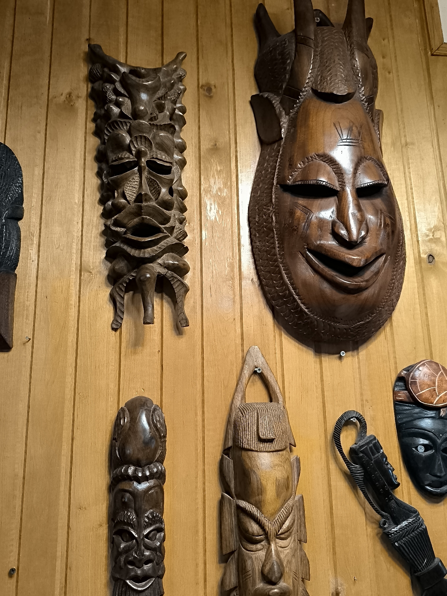 The image displays a collection of carved wooden masks and figures mounted on a light-colored wooden paneled wall. There are at least six distinct items visible. On the left, a tall, dark carved figure with a segmented body and a face is mounted vertically. To its right, a long, intricately carved dark brown mask with multiple faces and abstract elements is hung vertically. Further to the right, a large, reddish-brown wooden mask with a wide, smiling mouth and prominent features is centrally positioned. Below this large mask and to its left, a vertical carved figure with a head resembling a bird or fish on top of a face is displayed. To the right of this figure, another vertical mask, lighter in color and with a more angular design, is shown. To the far right, a black mask with a patterned circular element on top and what appears to be a carved handle or attachment extending downwards is visible. The wood grain is evident on the wall, and some of the masks show signs of age or wear. The lighting suggests an indoor setting. The image is a close-up shot of these artifacts. This scene is located in the village of Žiaukos, Lithuania, within a residential or decorative context.