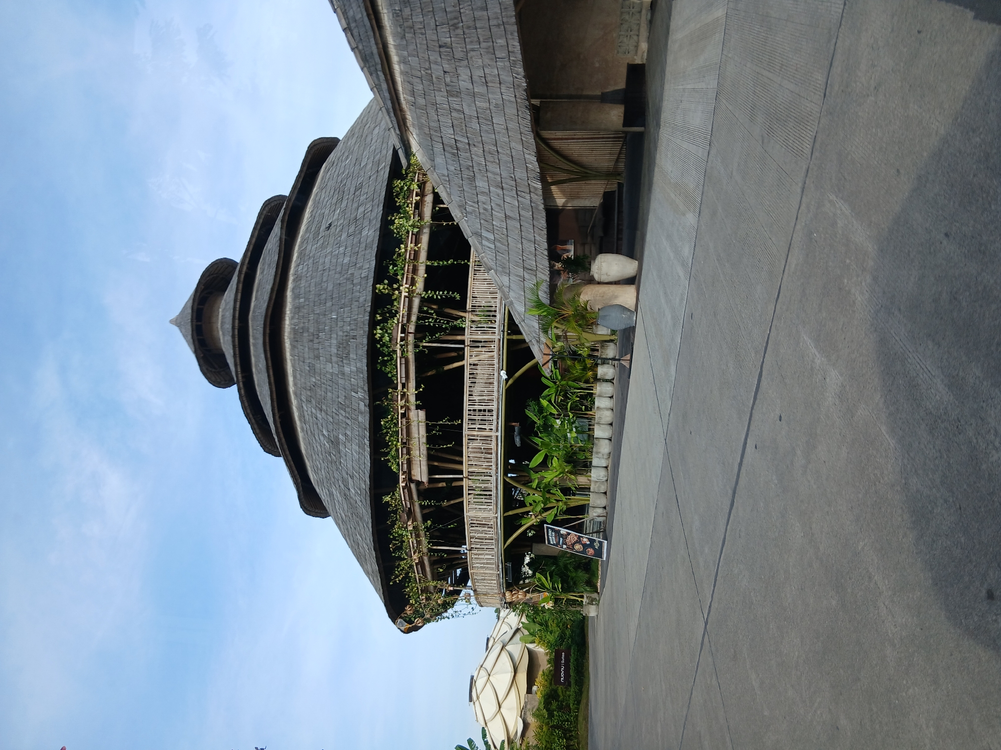This image depicts a unique architectural structure with a multi-tiered, thatched roof, likely a restaurant or resort building, located in the city of Kediri, Indonesia. The building features a circular main section with a wide, open-air balcony adorned with lush green vines. The balcony is supported by a bamboo structure. To the right of the main building, another section with a sloping thatched roof is visible, with an entrance area constructed from bamboo and concrete. In the foreground, a paved area with visible expansion joints suggests a public space or parking lot. Several large potted plants and decorative urns are placed near the entrance. A sign with a menu or advertisement stands near the plants. In the background, to the left, other dome-shaped structures with white, textured roofs are partially visible, hinting at a larger complex. The sky above is clear with scattered clouds. The overall scene is well-lit, indicating daytime. There are no people visible in the frame.