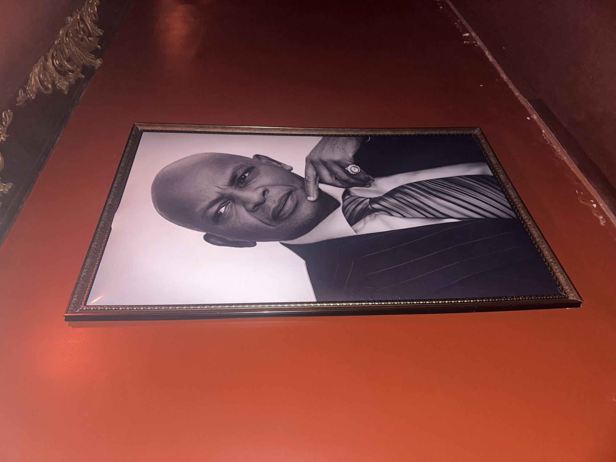 A framed black and white portrait of a man is mounted on a bright orange wall. The man is bald, wearing a pinstripe suit, white shirt, and a striped tie. He has his right index finger resting on his chin and is wearing a large ring on his right hand. The portrait is positioned at an angle, with the top right corner slightly higher than the bottom left. Above the portrait, a dark, ornate decorative molding is visible against a darker red wall. The lower portion of the image shows a dark red wall with a textured, uneven surface and some white marks near the floor, suggesting a corridor or hallway. The scene is likely an interior space within Abuja, Nigeria, possibly a hotel lobby, restaurant, or private residence given the decor.
