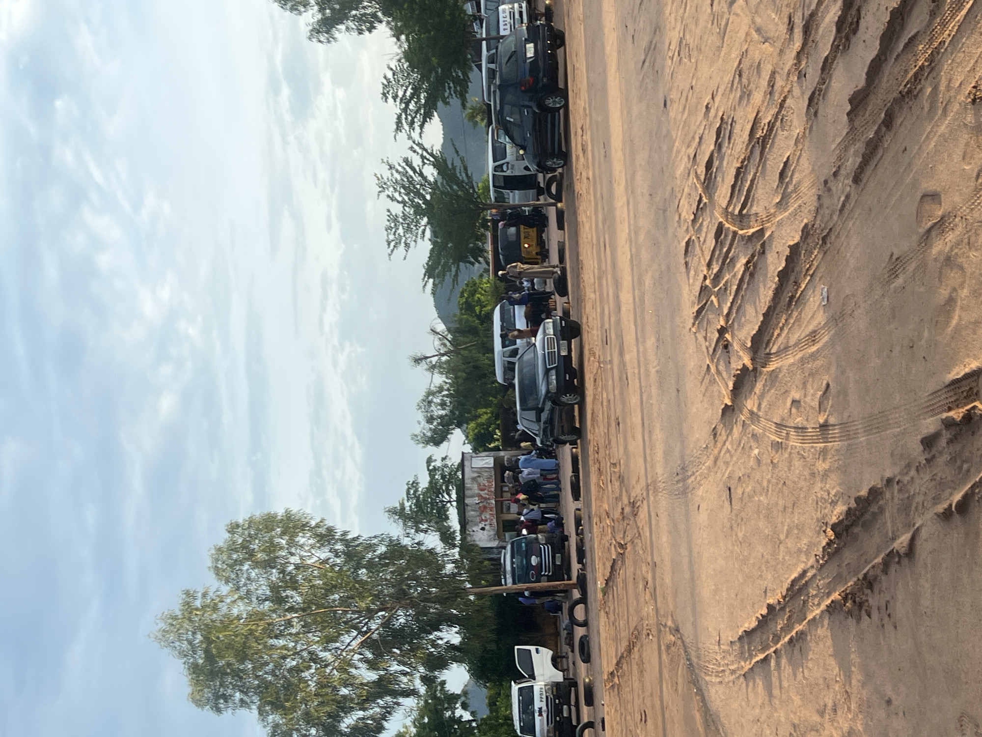 This image depicts an outdoor scene in Gbaza, Nigeria, characterized by an unpaved, reddish-brown dirt or sand lot, heavily marked with numerous vehicle tire tracks and ruts. The sky is light blue with scattered white clouds, suggesting daylight.
In the mid-ground, a small, single-story building with a weathered facade is visible. A faded sign on its front partially reads "T.Y.C" and "THOMAS OFFICE". Several individuals, approximately 15-20, are gathered near this building and among various parked vehicles.
To the left, a prominent tree with green foliage stands. Parked nearby is a white van with its side door open, displaying a license plate partially visible as "PP070". A dark-colored sedan is parked further to the left. In the central mid-ground, a black passenger van is parked.
Towards the right, multiple white vans are visible, with one having its side door open. A dark-colored SUV or crossover vehicle is parked on the right foreground. Further in the mid-right, a yellow tricycle (keke napep) is partially obscured by other vehicles. One individual in the mid-right is wearing light-colored traditional robes.
Several discarded or loose vehicle tires are scattered on the ground in the vicinity of the parked vehicles and the building. The background features distant green hills or mountains under the cloudy sky. The lighting suggests a natural daylight setting, possibly early morning or late afternoon, with soft shadows. The overall scene indicates an active transport or gathering point.