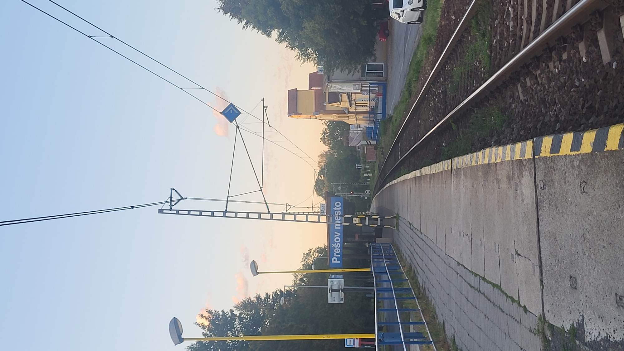 This is a full shot of a train station platform in Prešov, Slovakia. The platform is made of concrete slabs and has a yellow and black striped safety line along the edge. There are train tracks running alongside the platform. On the platform, there is a blue sign that says "Prešov mesto" which translates to "Prešov city". There are also lampposts, a ladder-like structure with wires, and some trees in the background. A few buildings are visible in the distance. The sky is clear and light blue, with some pinkish clouds. The scene appears to be during the day. There are no people visible in the image.