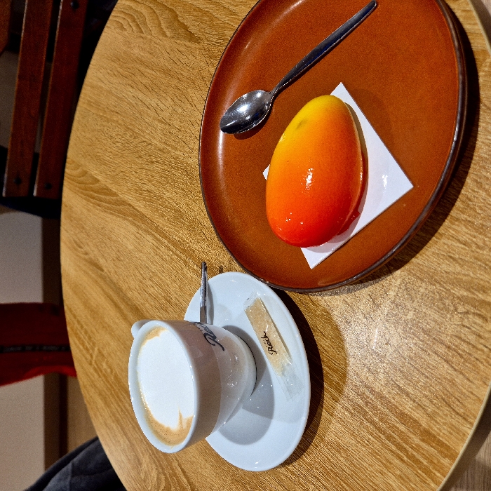 A top-down shot captures a wooden table setting. A glossy, egg-shaped dessert, shaded from orange to red, sits on a white napkin on a brown plate, accompanied by a spoon. Beside it, a white cup of coffee rests on a matching saucer with a sugar packet. A dark garment is subtly visible near the cup. A partial view of a wooden chair appears in the background.