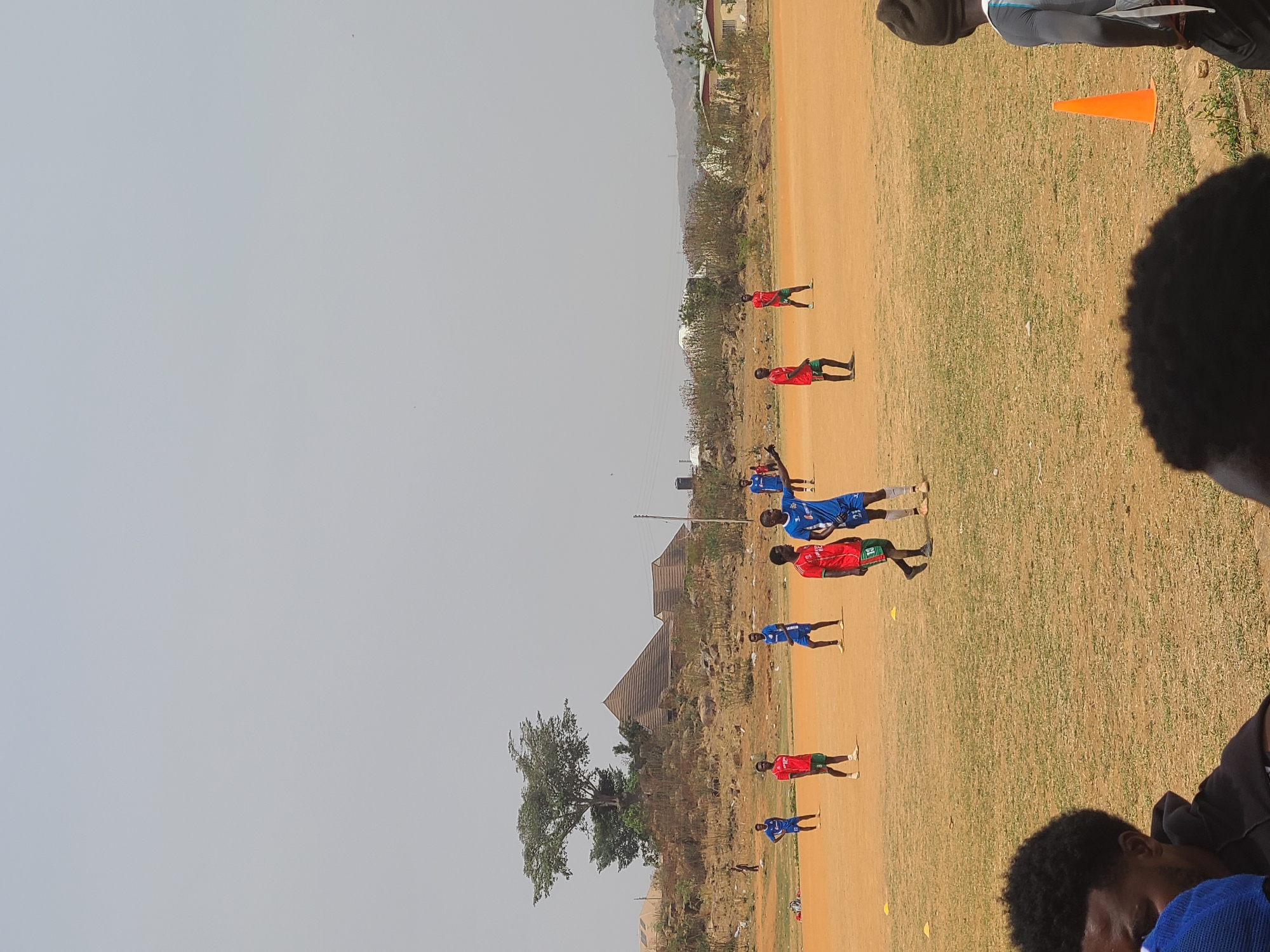 The image depicts a soccer training session in Barikin Jirga Summa, Nigeria. Players in red and blue jerseys are positioned on a dirt field with scattered grass. There are yellow cones placed along the field. In the background, there are trees, buildings with corrugated metal roofs, and hills. The sky is clear. Several people, their faces partially visible, are observing the training session from the sidelines.