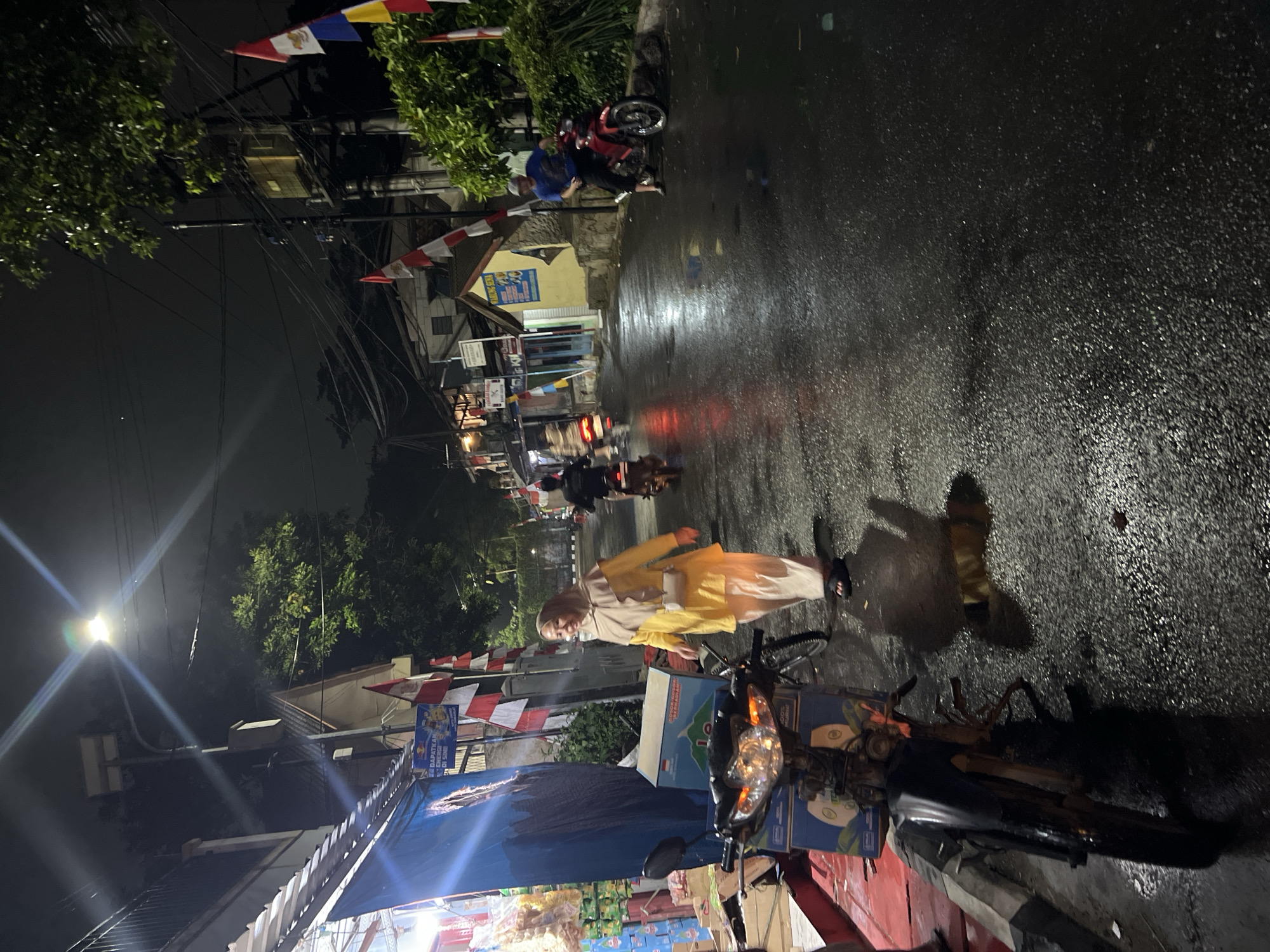 A nighttime street scene in Citeureup, Indonesia, illuminated by streetlights and the glow of shops. The asphalt road is wet and reflects the lights, indicating recent rain. Several people are present, including a woman in a yellow hijab and outfit walking on the sidewalk, and a man in a blue t-shirt sitting on a red motorcycle. Other individuals are seen on motorcycles in the background, moving along the street. Buildings line the right side of the road, with flags prominently displayed outside some of them. On the left, a storefront is visible with goods displayed on shelves. A parked motorcycle is in the foreground on the left, with boxes stacked on it. The overall atmosphere is that of a busy, illuminated street at night, possibly a commercial area, with ongoing traffic and pedestrian activity.