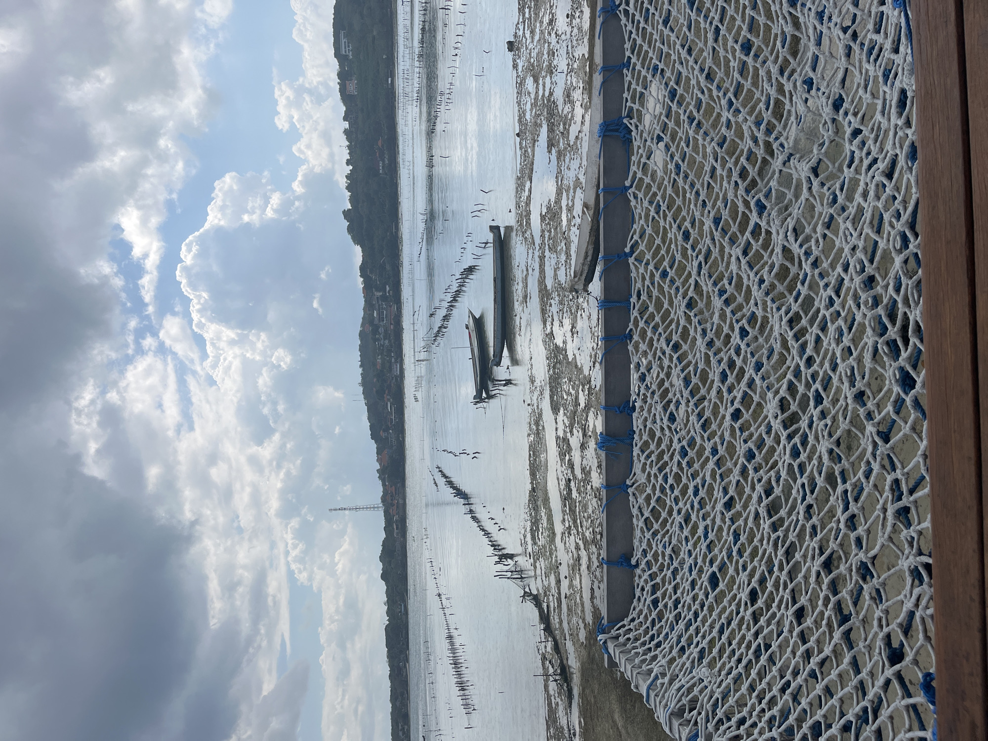 This image depicts a serene coastal scene in Nusa Penida, Indonesia, likely a shallow bay or estuary used for aquaculture. In the foreground, a white net hammock with blue rope accents is stretched over a wooden frame, hinting at a relaxing vantage point. Beyond the hammock, the water is calm and shallow, revealing a muddy seabed dotted with numerous wooden posts arranged in neat rows, indicative of seaweed or fish farming. Several small boats, some partially submerged and others floating gently, are scattered across the water. In the middle ground, a line of taller wooden structures extends into the water, possibly part of the farming infrastructure. The background features a lush, green hillside dotted with houses and buildings, suggesting a coastal village. A tall communication tower stands prominently on the hill. The sky is a mix of dramatic, cloudy formations and patches of bright blue, with the sunlight reflecting off the water's surface. The overall atmosphere is peaceful and tranquil, capturing the essence of a coastal working landscape.