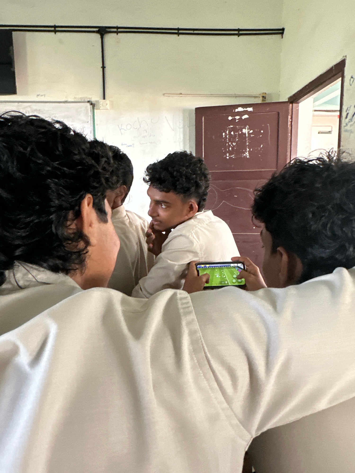In Malappuram, India, within a room, a group of young men in light-colored shirts are gathered. One young man in the foreground is holding a smartphone, actively engaged with a game displaying a green field with circular elements. His attention is directed towards the screen. Another young man to his left is looking towards the phone, possibly observing the game. In the background, a brown door is ajar, revealing a glimpse of another room. The walls are light-colored, with some electrical conduits running across them. Whiteboards are visible on one wall, with faint writing on them. The overall atmosphere suggests a casual gathering, possibly during a break.