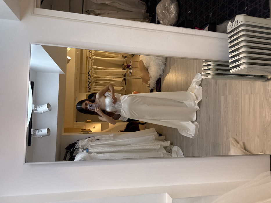 A woman in a white wedding dress takes a mirror selfie in a bridal shop. Her phone covers her face. The dress has a sparkly, corset-style bodice and a long, flowing skirt. Racks of wedding dresses are behind her. The store has spotlights on the ceiling, and a white portable radiator stands against the wall near the mirror. A second person is in the background next to her.