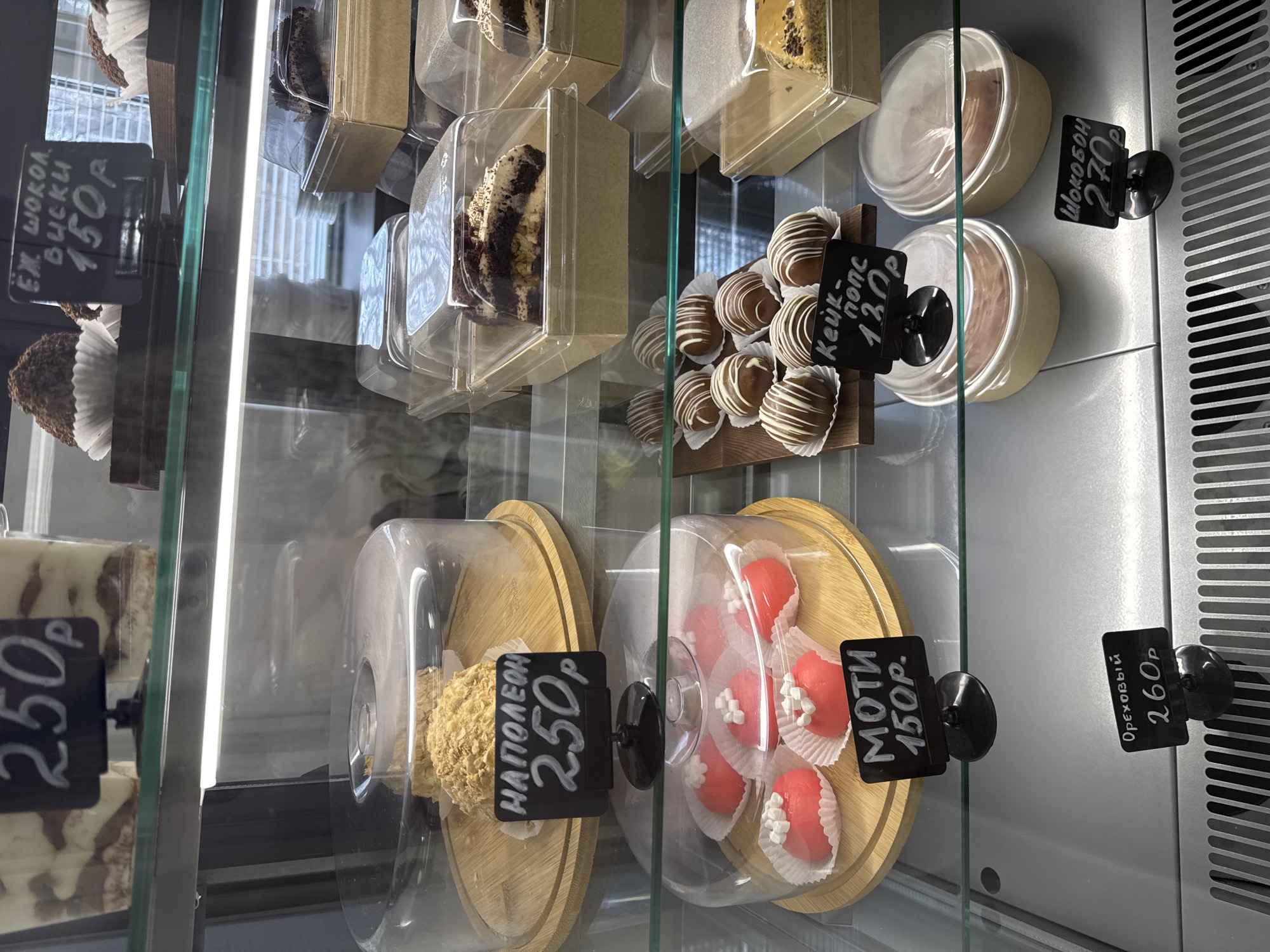 This is a close-up photograph of a pastry display case, likely in a store in Chaikovsky, Russia. The case contains various cakes and desserts, each with a price tag in Russian rubles. Visible items include:
* Chocolate hedgehog cakes (top left, priced at 150 rubles)
* Sliced cakes in clear plastic containers (center top)
* Chocolate-covered cake pops (middle right, priced at 120 rubles)
* Chocolate pudding or mousse in small containers (top right, priced at 270 rubles)
* "Napoleon" cake under a dome (bottom left, priced at 250 rubles)
* "Mochi" cakes, also under a dome (bottom center, priced at 150 rubles)
* "Nutty" cake/dessert (bottom right, priced at 260 rubles).
The display case has glass shelves and a metal frame with ventilation slots on the right side. The interior is brightly lit. The photo appears to have been taken indoors.
