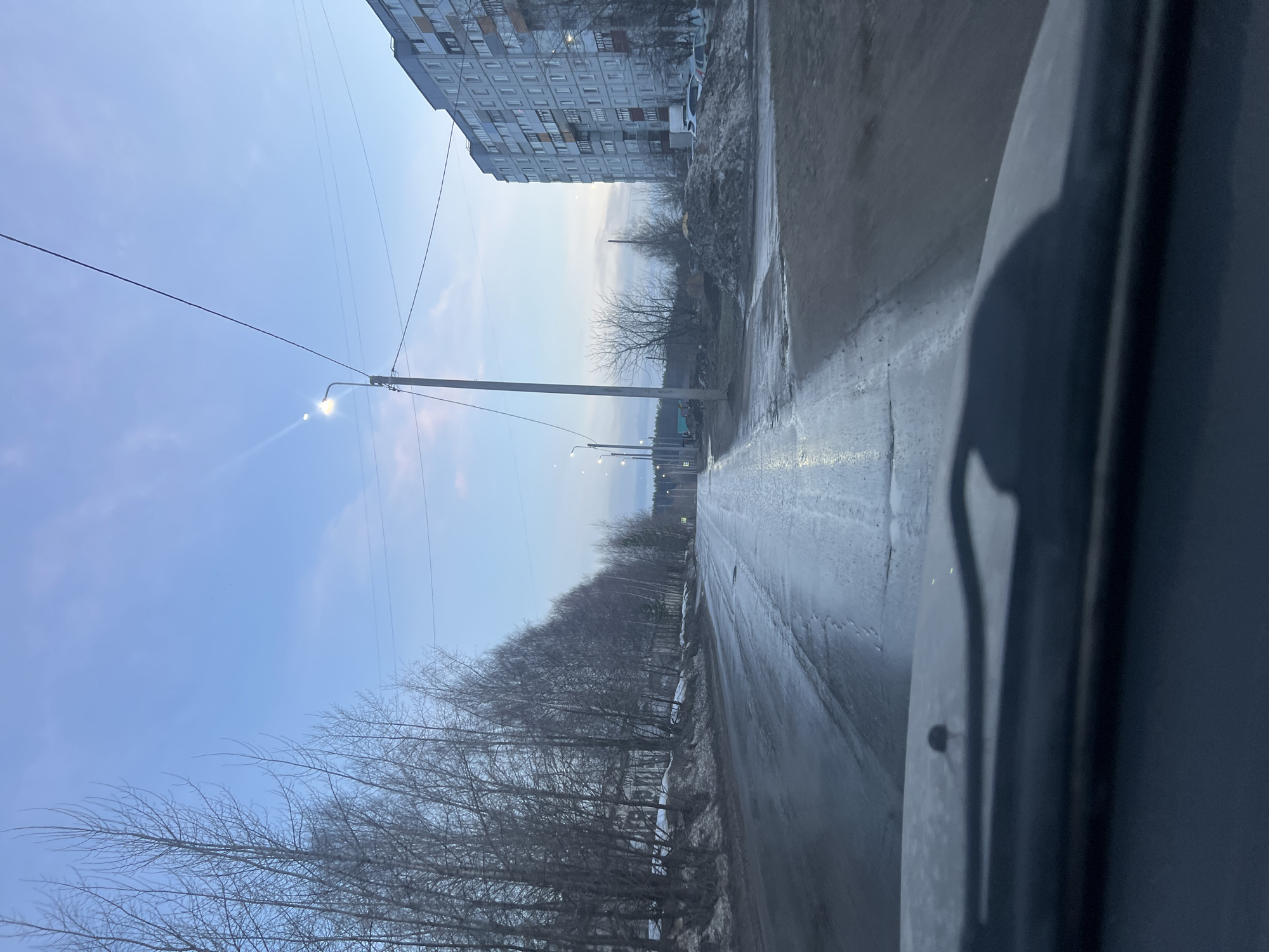 The image shows a street scene in what appears to be the city of Chaikovsky, Russia. A multi-story apartment building is on the left side of the road. Power lines and lampposts are visible. Bare trees line the opposite side of the road, which is wet and appears to have patches of snow or ice. The photo appears to be taken from inside a vehicle, with part of the dashboard visible in the foreground. The sky is light blue with some cloud cover. No people are visible.