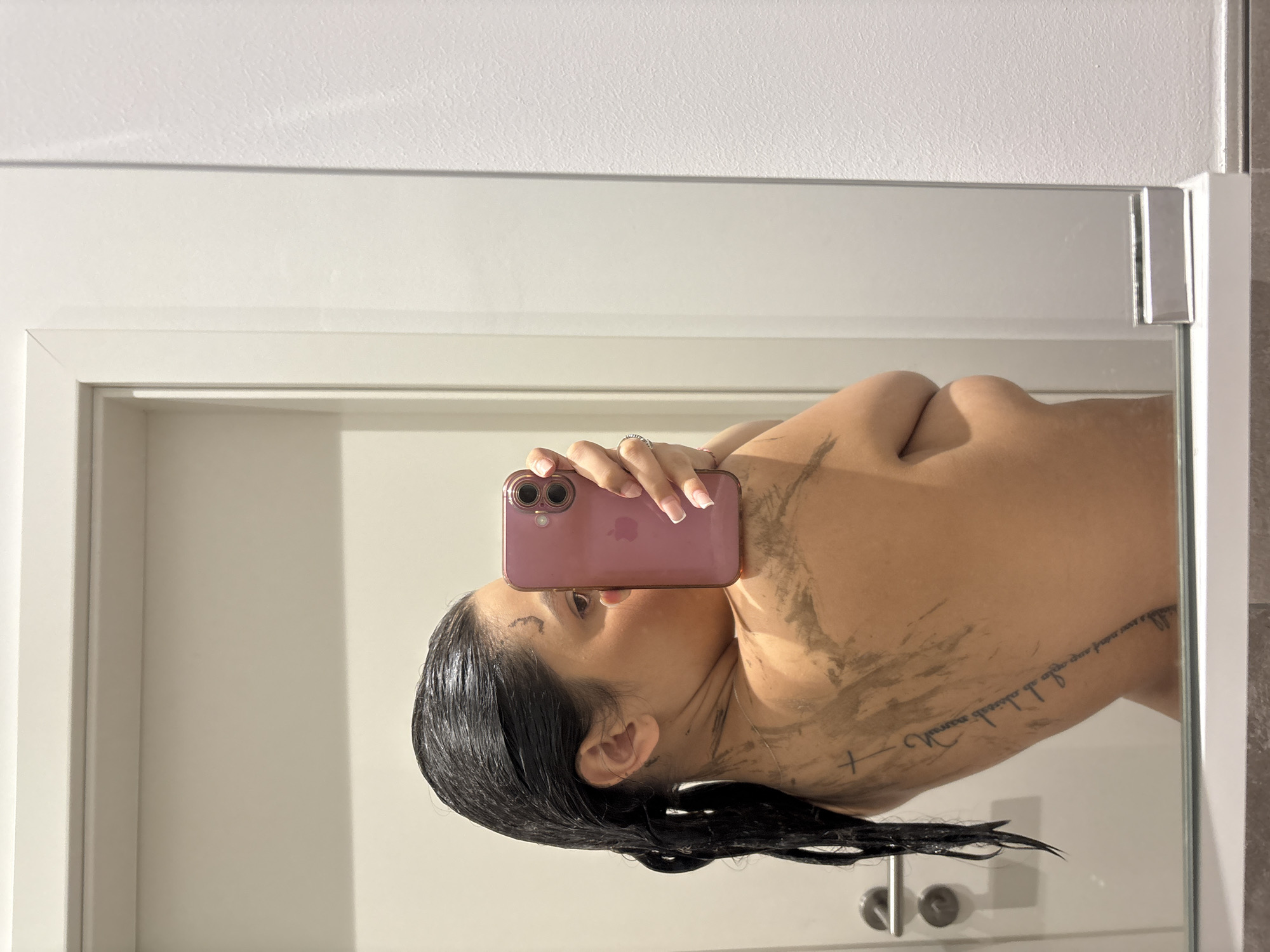 The image, captured in Mönchengladbach, Germany, depicts a woman taking a mirror selfie indoors. The subject is seen from the mid-torso up, facing her right, with her back partially presented towards the viewer's left. Her dark hair appears wet, slicked back, and partially clumped together. Her skin, particularly on her upper back and left shoulder, is covered in dark, streaky, brownish-grey residue consistent with mud or a clay application.
She holds a pink-cased Apple iPhone horizontally with her right hand, obscuring the majority of her face; only her right eye and part of her right cheek are visible. A silver-toned ring is present on the fourth finger of her right hand, and her fingernails exhibit light-colored polish. Tattoos are visible: a thin, vertical line of script along her left rib cage/back, terminating with a small cross symbol near her left shoulder blade, and a small stylized symbol or script on her right forehead/temple area. A faint dark marking is also present behind her left ear. A thin, gold-colored necklace chain is discernible around her neck.
The background reflected in the mirror consists primarily of a plain off-white wall. To the left of the reflection, a white door with two metallic, cylindrical door handles is visible. To the right of the mirror's reflection, a textured white wall is present. The mirror itself features a white frame, and its lower edge reveals a narrow, grey, tiled ledge.