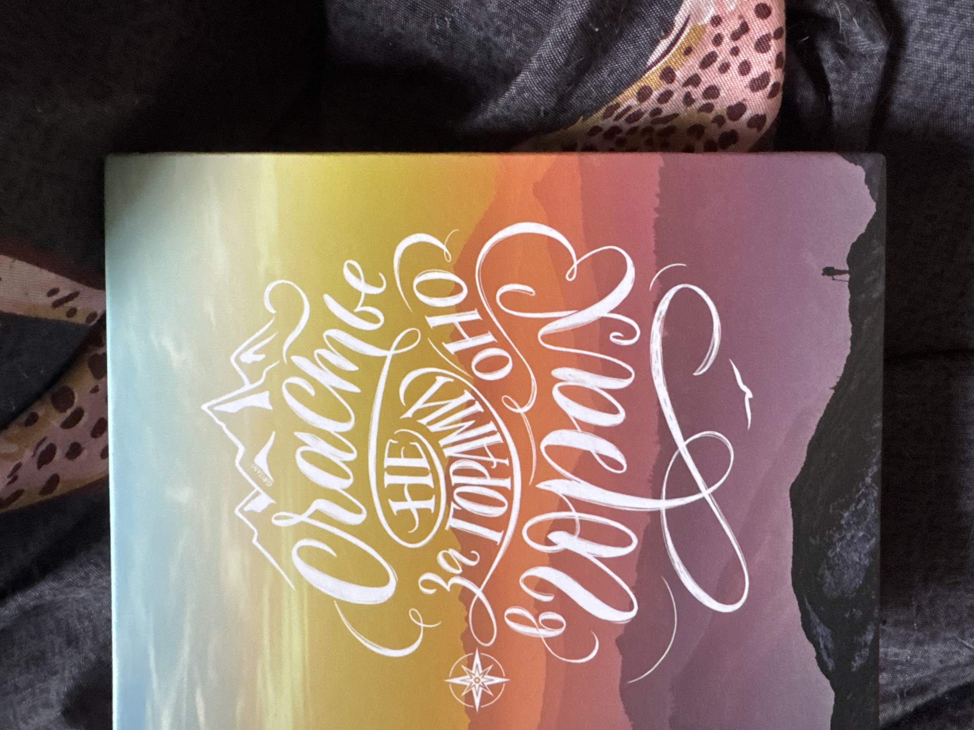 The image shows the cover of a notebook or book. The background features a gradient of colors, transitioning from a light yellow and green on the left to orange and purple on the right, resembling a sunset or sunrise over a mountainous landscape. A dark silhouette of a mountain or cliff is visible on the far right, with a tiny silhouette of a person standing on the edge.
Dominating the left and center of the cover is intricate white calligraphy. The script is stylized and decorative, forming words in Russian. The main phrase appears to be "Счастье не за горами," which translates to "Happiness is not far away." The calligraphy is overlaid on the colorful background and also incorporates mountain-like shapes within the lettering, particularly in the word "Счастье" (happiness). A small compass rose symbol is subtly placed at the bottom center of the text.
The notebook is placed on a dark grey textured fabric, possibly a blanket or clothing, with a patterned fabric with brown spots and a pinkish hue peeking in from the top left and top right corners. The overall mood conveyed by the image is one of inspiration, optimism, and adventure, with the imagery of mountains and the call to find happiness suggesting a journey or a positive outlook. The lighting suggests it could be daytime, perhaps during the golden hour due to the warm colors in the background. There are no specific weather cues visible, but the serene landscape implies pleasant conditions.