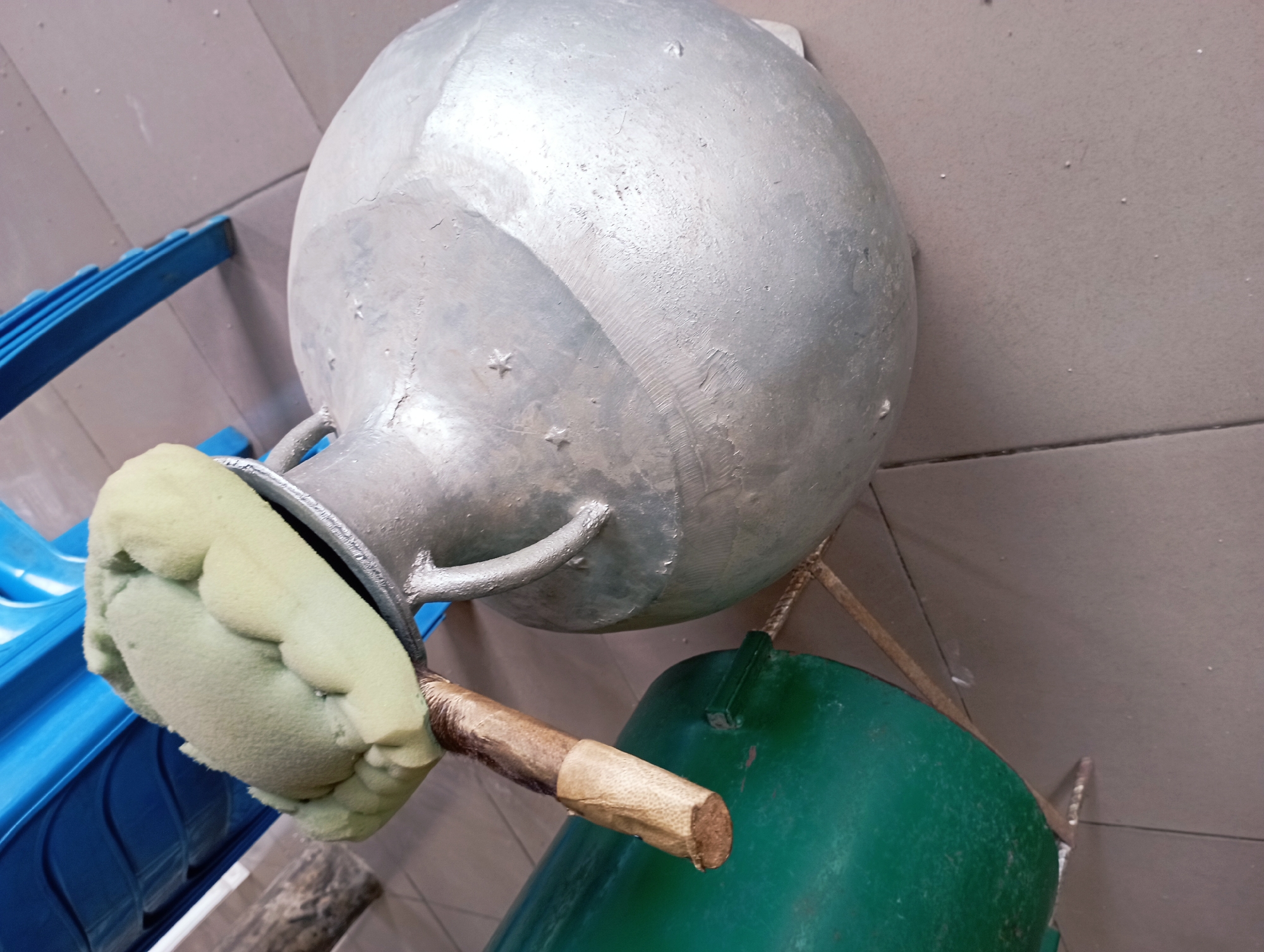 A close-up, high-angle shot captures a light gray, bulbous metal pot with two handles. The pot sits on a tiled floor, likely in Jos, Nigeria. A wooden stick, wrapped in light brown tape, extends from a green container positioned behind the pot. A pale green, foamy material is attached to the end of the stick, resting on the rim of the metal pot. A portion of a blue plastic chair is visible to the right of the scene. The overall impression is one of an outdoor or utilitarian setting.