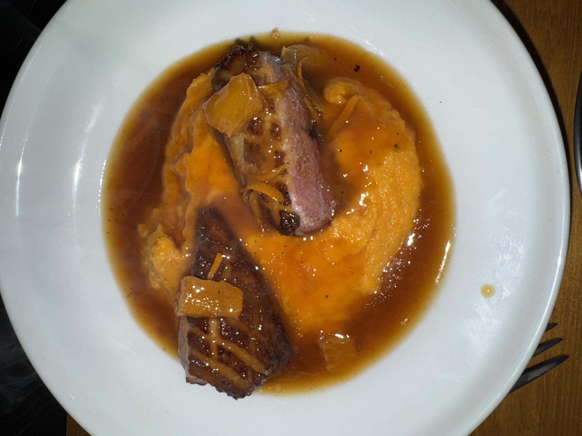 A close-up, overhead view captures a hot meal presented on a white, slightly oval plate. The central focus is a dish featuring two pieces of seared duck breast, displaying a crisped, dark-brown skin and a pink, medium-rare interior. These are situated within a pool of rich, amber-brown sauce, which also envelops a large portion of vibrant orange purée, consistent with sweet potato or pumpkin. The duck and sauce are garnished with pieces of translucent orange peel or zest. Wisps of steam are faintly visible rising from the dish, indicating its temperature. Small, dark particulate matter, possibly ground pepper or herbs, is sparsely distributed across the sauce and purée. The white plate exhibits a subtle sheen and a small, isolated drop of the amber sauce on its rim towards the bottom-right. The plate rests on a textured, light brown wooden surface. A dark metal fork is partially visible underneath the plate on the lower-left. The background is uniformly dark and out of focus. No individuals are present in the frame, nor are any direct actions or interactions depicted. This scene is situated in what is indicated to be a location in Paris, France.