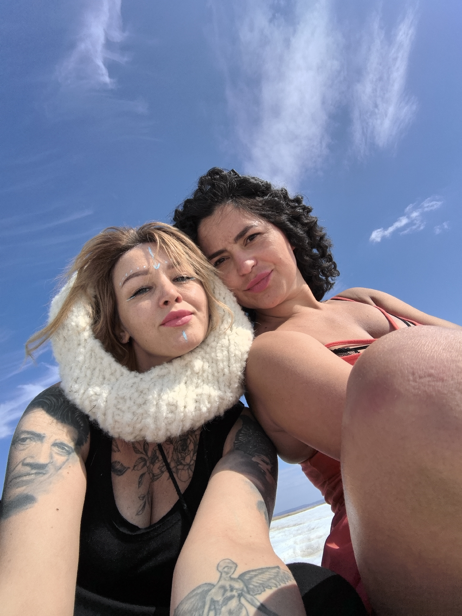 A low-angle selfie captures two women against a bright blue sky with scattered wispy white clouds. The foreground shows a woman with blonde hair, distinctive light blue face paint on her forehead, cheek, and chin, and pink lipstick. She wears a thick, off-white knitted scarf or hood around her neck and a black tank top, revealing numerous tattoos on her arms and chest, including a portrait on her left bicep, an angelic figure on her left forearm, and floral designs on her chest. She looks directly at the camera with a calm expression.
Partially visible behind her to the right is another woman with dark, curly hair, smiling softly. She is wearing a reddish-orange top, possibly a swimsuit. Her arm and bent leg are visible, suggesting she is seated or lounging.
The sky occupies the majority of the upper frame, indicating an expansive outdoor setting. Below the subjects, a narrow strip of a vast, flat, light-colored landscape is visible, possibly snow, sand, or salt flats. The bright light suggests it is daytime, likely under a clear, sunny sky. Both women appear relaxed and content. There is no visible text in the image. The scene is set in an open environment, consistent with a remote location in Noviy, Russia.
