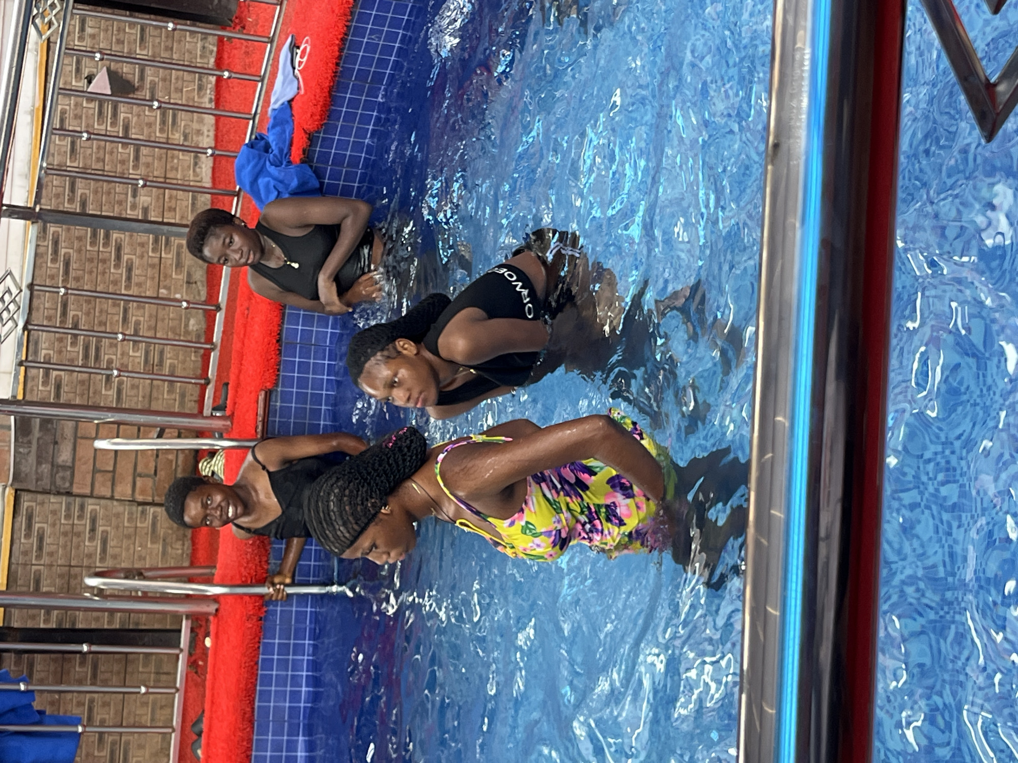 Four young women are in a swimming pool. One woman in the foreground on the left is wearing a yellow and green floral swimsuit. To her right and slightly behind her, another woman is wearing a black tank top. In the background, two more women are in the pool. One on the left is wearing a black tank top and smiling. The woman on the right is also wearing a black tank top. The pool has blue tiled walls and is filled with clear blue water. There are metal railings and a red mat surrounding the pool. The scene appears to be at a recreational facility in Obosi, Nigeria.