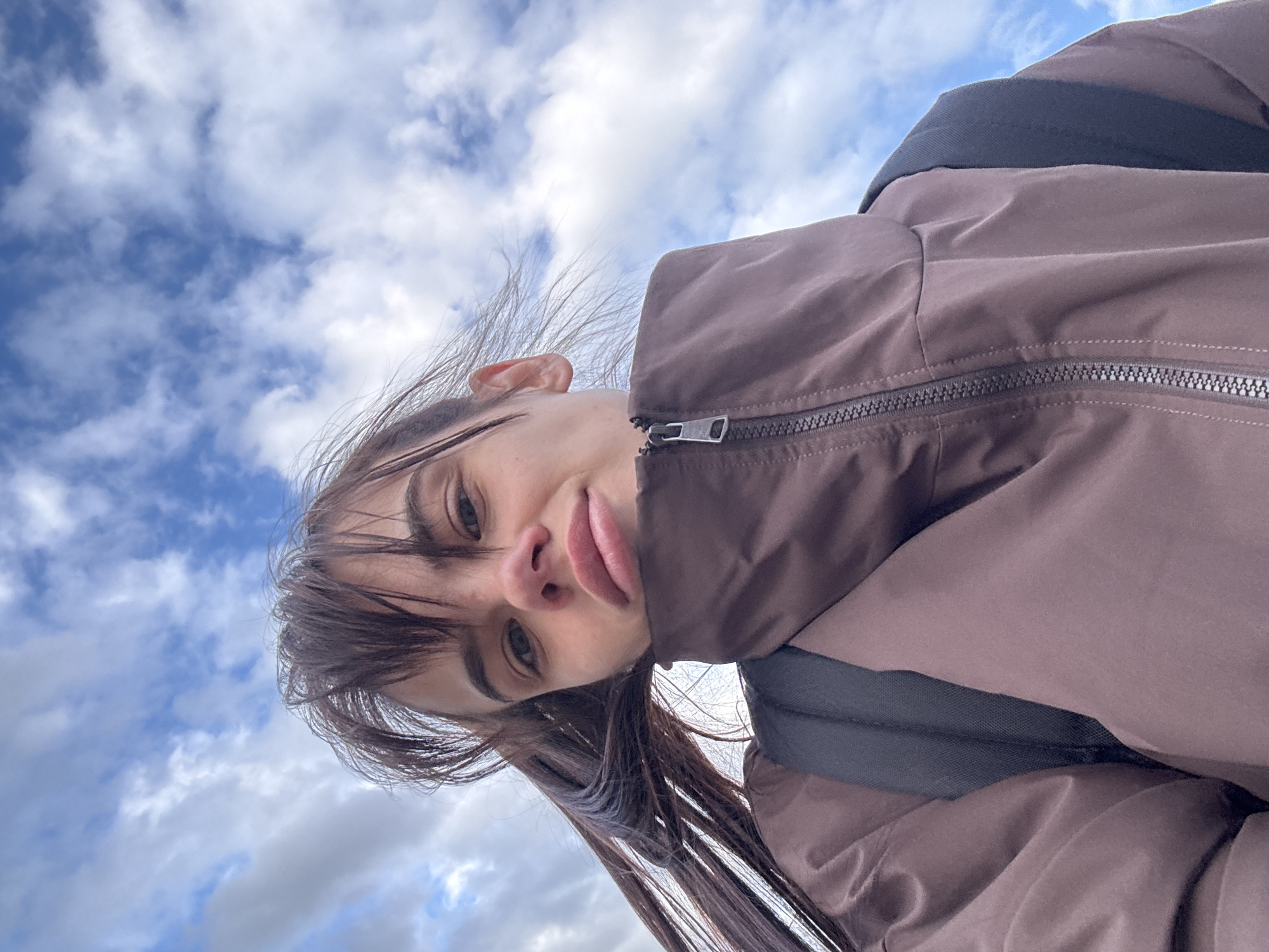 A low-angle, close-up selfie shows a young female outdoors, potentially in Rostov-on-Don, Russia. Her face is tilted, and she looks directly at the camera with a playful expression, her tongue slightly protruding. She has dark hair, including bangs, and light eyes.
She is wearing a brown or dark beige jacket with a prominent black zipper down the front. A black strap, likely from a backpack, is visible over her right shoulder, suggesting she is carrying a bag. The background is a bright daytime sky filled with large, white, fluffy clouds against a light blue expanse. The natural light is soft, indicating an overcast but clear day. No specific location cues other than the sky are visible within the image itself. There is no visible text.