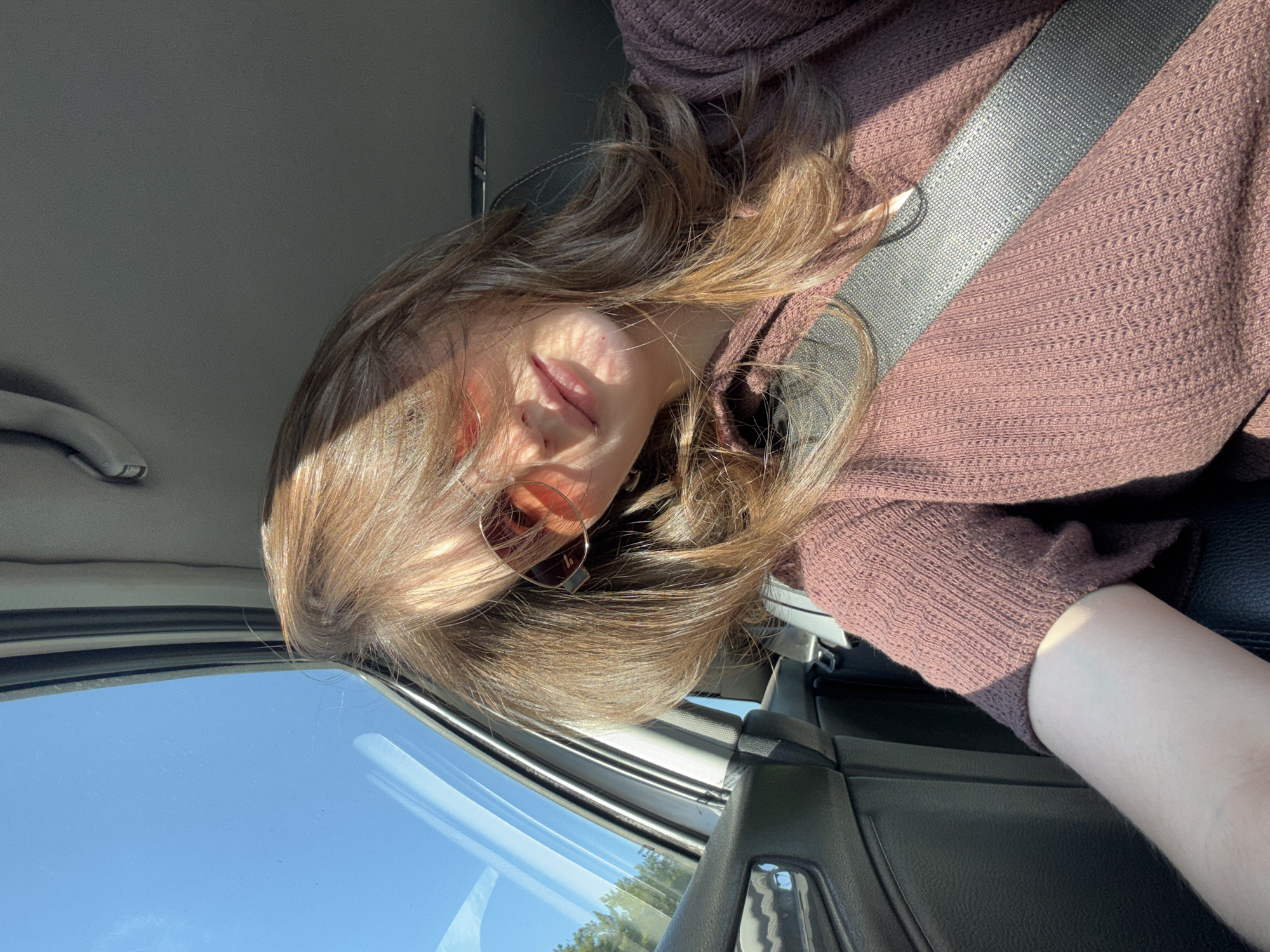 A woman is sitting in a car. She is wearing a brown sweater and a seatbelt. The window is visible, with blue sky showing through. The car's interior is predominantly black and gray. The location is unknown and cannot be inferred from the image.