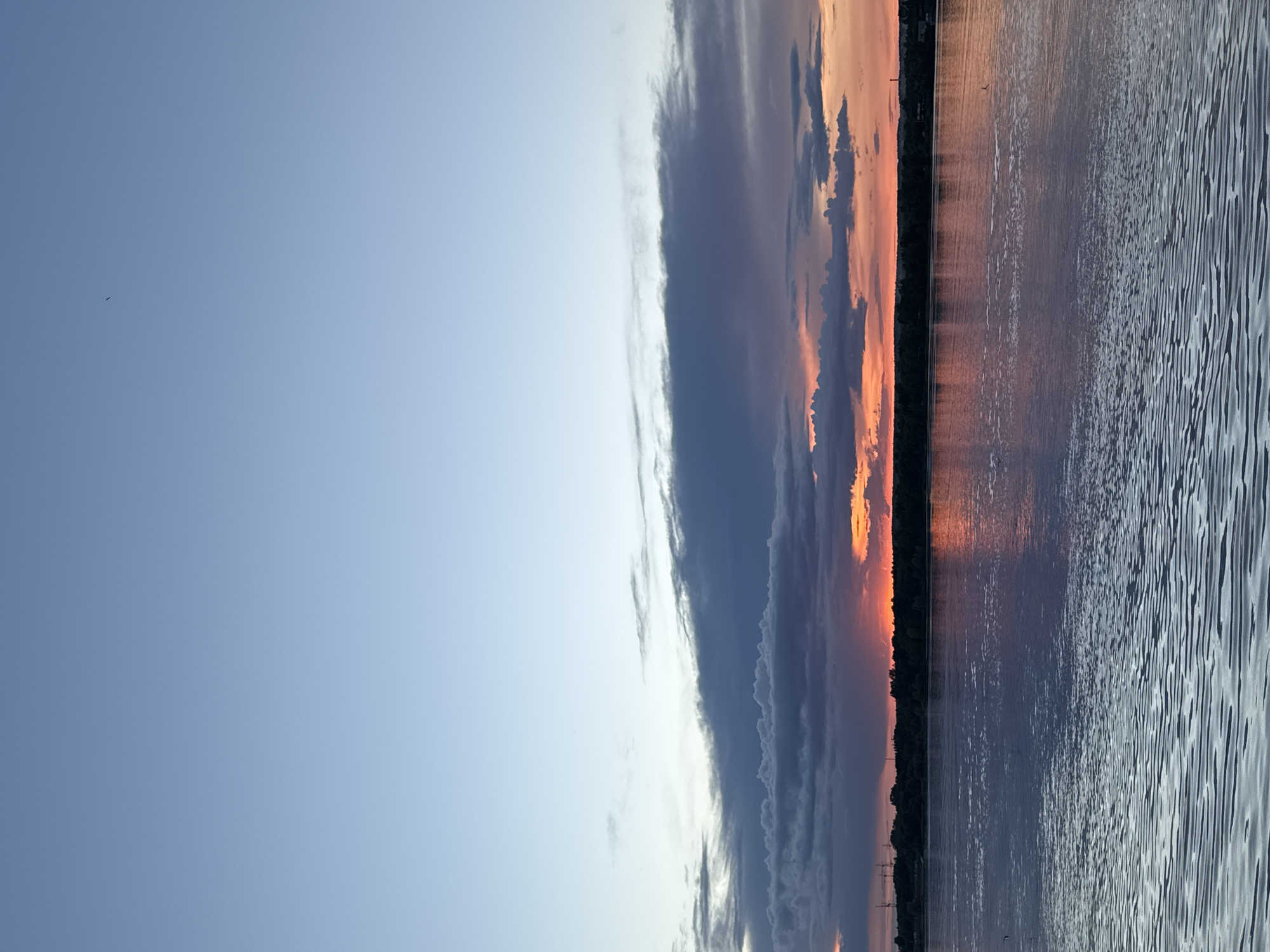 The image displays a picturesque sunset scene over a large body of water in Perm, Russia. The composition is horizontally divided between the sky and the water, with a thin line of land marking the distant horizon.
The upper half of the image is dominated by the sky, which transitions from a clear, light blue on the left to dramatic sunset colors on the right. A prominent, dark band of clouds stretches horizontally across the sky, separating the clearer blue from the vibrant hues of dusk. To the right, the clouds are illuminated by the setting sun, glowing in shades of orange, pink, and red.
Below the colorful sky, a dark, thin line of what appears to be a distant shoreline or treeline is silhouetted against the bright light of the sunset.
The lower half of the image is an expanse of water, reflecting the sky's dynamic colors. The surface of the water shows ripples and textures, particularly noticeable towards the right side, indicating gentle movement. The reflections beautifully mirror the dramatic clouds and the warm glow of the sunset, creating a harmonious and immersive natural vista.
No people or explicit man-made objects are visibly present, focusing entirely on the natural landscape. The time of day is clearly sunset or dusk, characterized by the intense and warm coloration of the sky and its reflections. The overall atmosphere is serene and captivating. No visible text is present in the image.