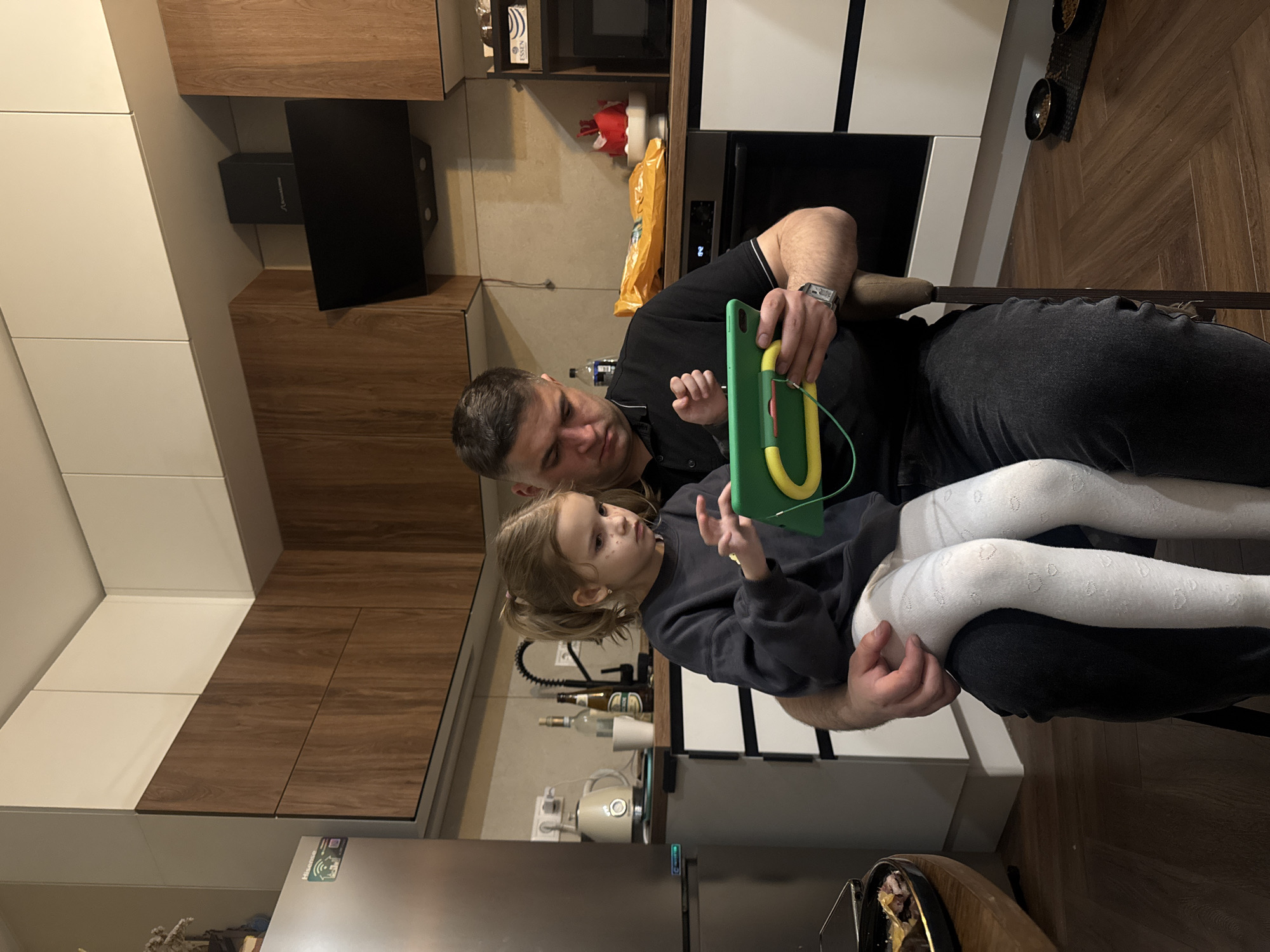 The photograph shows a man and a young girl indoors, presumably in a kitchen. The man is seated, wearing a black shirt and dark pants, with the girl sitting on his lap. The girl is wearing a dark gray sweater and white leggings. They are both looking at a green tablet device, which the man is holding. Kitchen cabinets are visible in the background, with a combination of white and wood-grain finishes. There is a microwave and other kitchen appliances in the background. Based on the items present and the modern kitchen design, the location appears to be a residential space. It is difficult to confirm with certainty that the location is Razvilka, Russia based only on the visual information provided in the image.