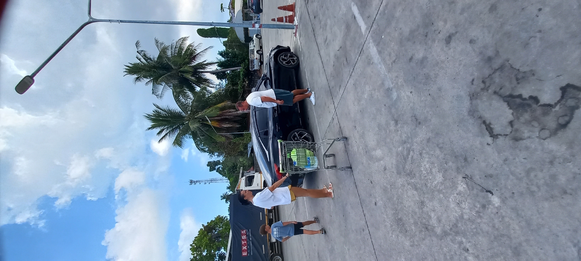 The image depicts an outdoor scene in a sunlit parking lot, likely associated with a supermarket in a tropical environment. The sky is bright blue with scattered white clouds, and several tall palm trees are prominent in the mid-ground, suggesting a warm climate like Moo 1, Thailand.
In the foreground, a black passenger car, possibly an SUV or hatchback, is parked on concrete pavement with its rear door open. A metal shopping cart, containing several light green plastic bottles, is positioned next to the car's open trunk area. Two adults are present, seemingly engaged in loading items. One adult male, wearing a white t-shirt and dark shorts, is leaning into the car's open rear. Another adult, dressed in a white t-shirt and yellow shorts or skirt, stands closer to the shopping cart, facing the car.
A child, wearing a blue t-shirt and dark shorts with "WSL" printed on it, is visible further back near a large black truck or trailer. The word "EXTRA" is clearly visible in white text on the side of this truck. In the background, beyond the truck, more greenery, a communication tower, and orange traffic cones can be seen. A tall street light pole with a long arm extends into the sky from the left side of the frame. The concrete ground of the parking lot shows various dark stains and markings. The overall scene portrays a routine activity of loading purchases into a vehicle on a bright, clear day.