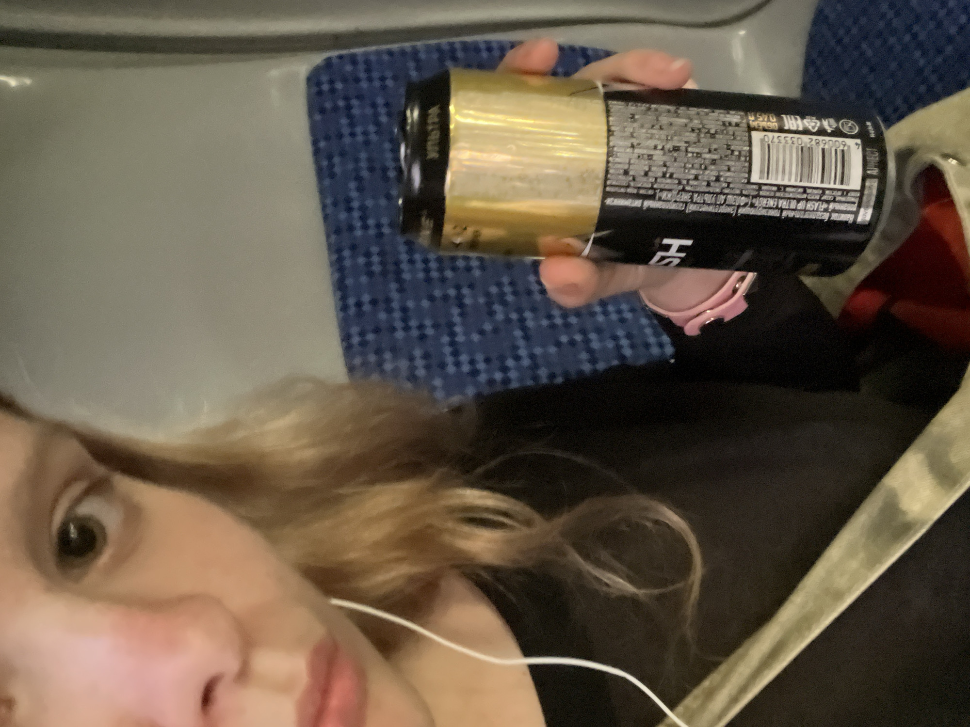 A close-up, slightly blurry, medium shot shows a person holding a black and gold can of energy drink. The person, whose face is partially visible in the upper left, has fair skin, light brown hair, and is wearing a black top. A white earphone wire is visible. The can, held in the person's right hand, has Russian text and a barcode on its black portion. The background features a blue patterned seat, likely in a public transportation setting. The image does not contain any specific identifiable landmarks or features of Kokkorevo, Russia.