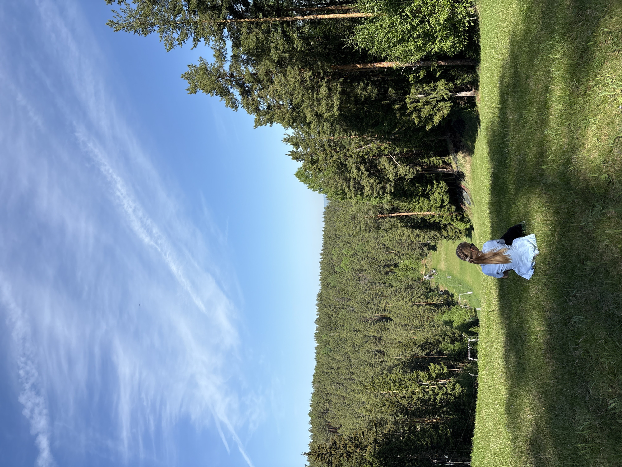 A girl is sitting on a grassy slope in Прикамский, Russia. The sky is blue with streaks of clouds. Dense forest is behind the slope.