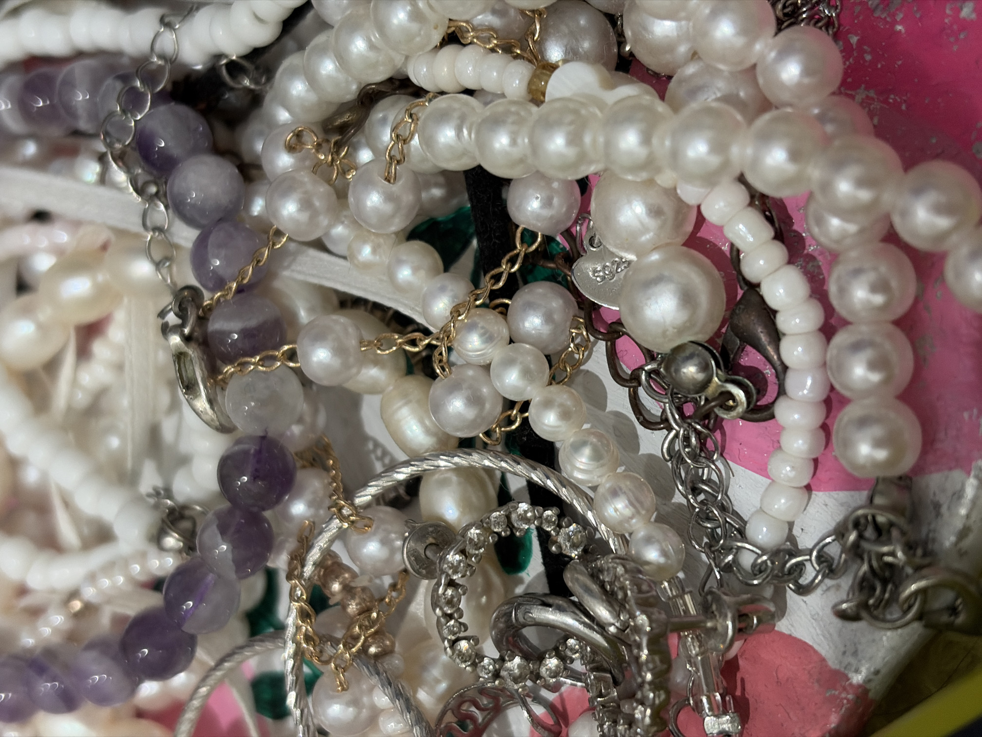 A close-up photograph shows a jumble of jewelry, primarily necklaces and bracelets, overlapping and entangled. The materials include white pearls, white beads, light purple amethyst beads, gold and silver-toned chains, and silver-toned metal settings, some with embedded clear stones. No people are visible. The background appears to be a surface painted with pink, white, and other colors. There is no indication of this image being taken in Izhevsk, Russia.