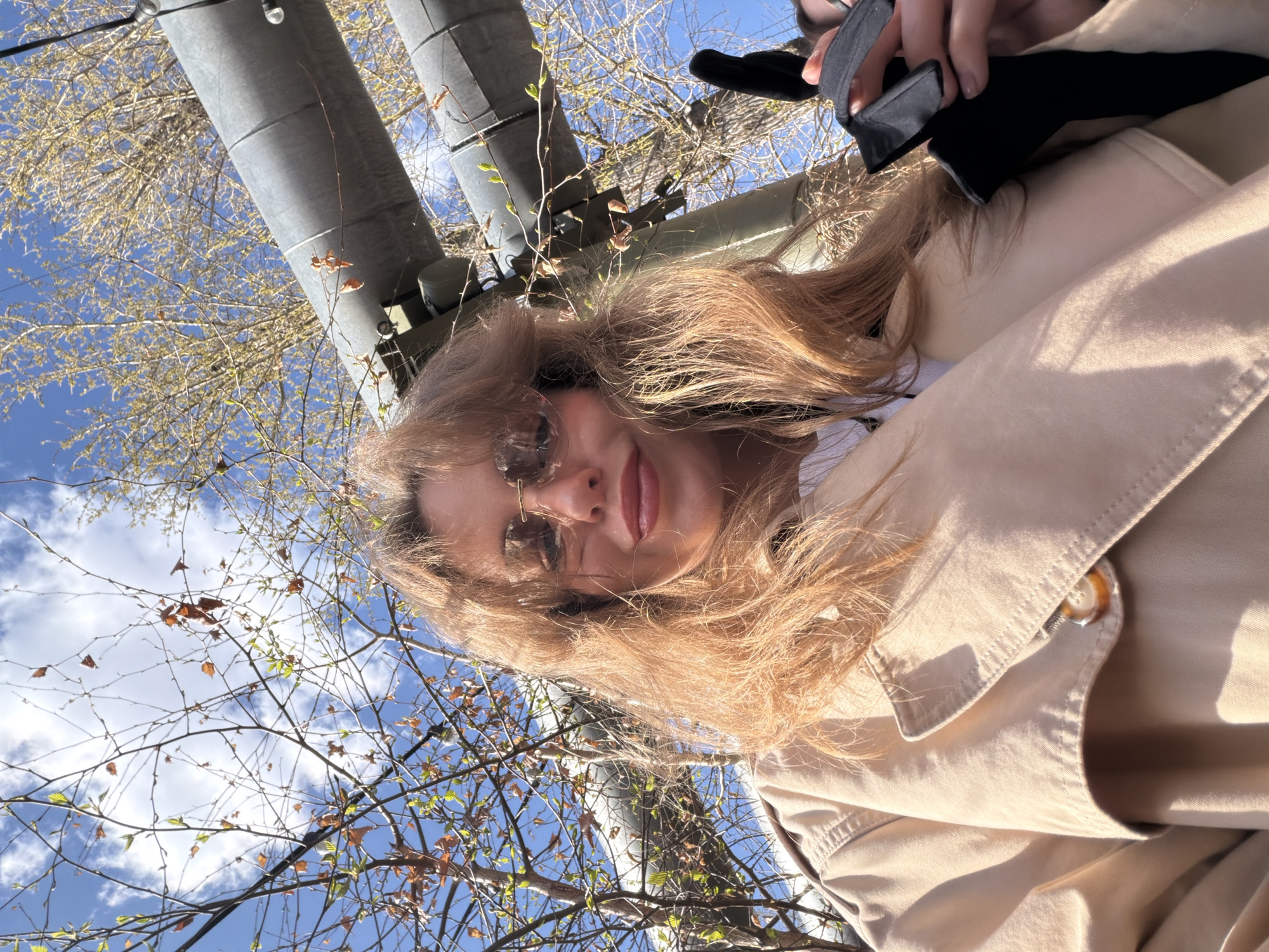 A woman with auburn hair and sunglasses is shown from an upward angle. She is wearing a beige trench coat. Above her is a blue sky with scattered clouds and tree branches with sparse foliage. Metal pipes and utility structures are also visible. This photo was taken in Moscow, Russia.
