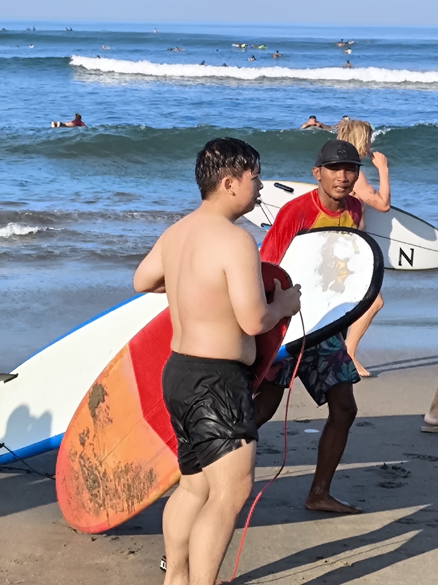 The image depicts a beach scene in North Kuta, Indonesia, with several people and surfboards. In the foreground, a shirtless man in black shorts carries a surfboard. Next to him, another man wearing a red and yellow rash guard, black baseball cap, and floral-patterned shorts carries a bodyboard. Further back, a person with blonde hair carries a white surfboard with the letter "N" on it. Several individuals are visible surfing in the ocean in the background. The scene suggests a surfing lesson or recreational activity on the beach.