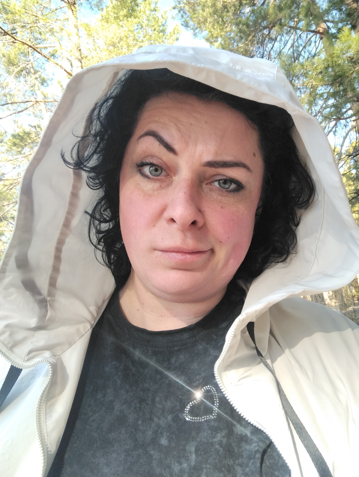 A woman is posing for a selfie in an outdoor environment with trees and foliage visible in the background. She is wearing a black t-shirt with a decorative heart design and a light-colored hooded jacket. The location is possibly a park or wooded area within the city of Votkinsk, Russia.