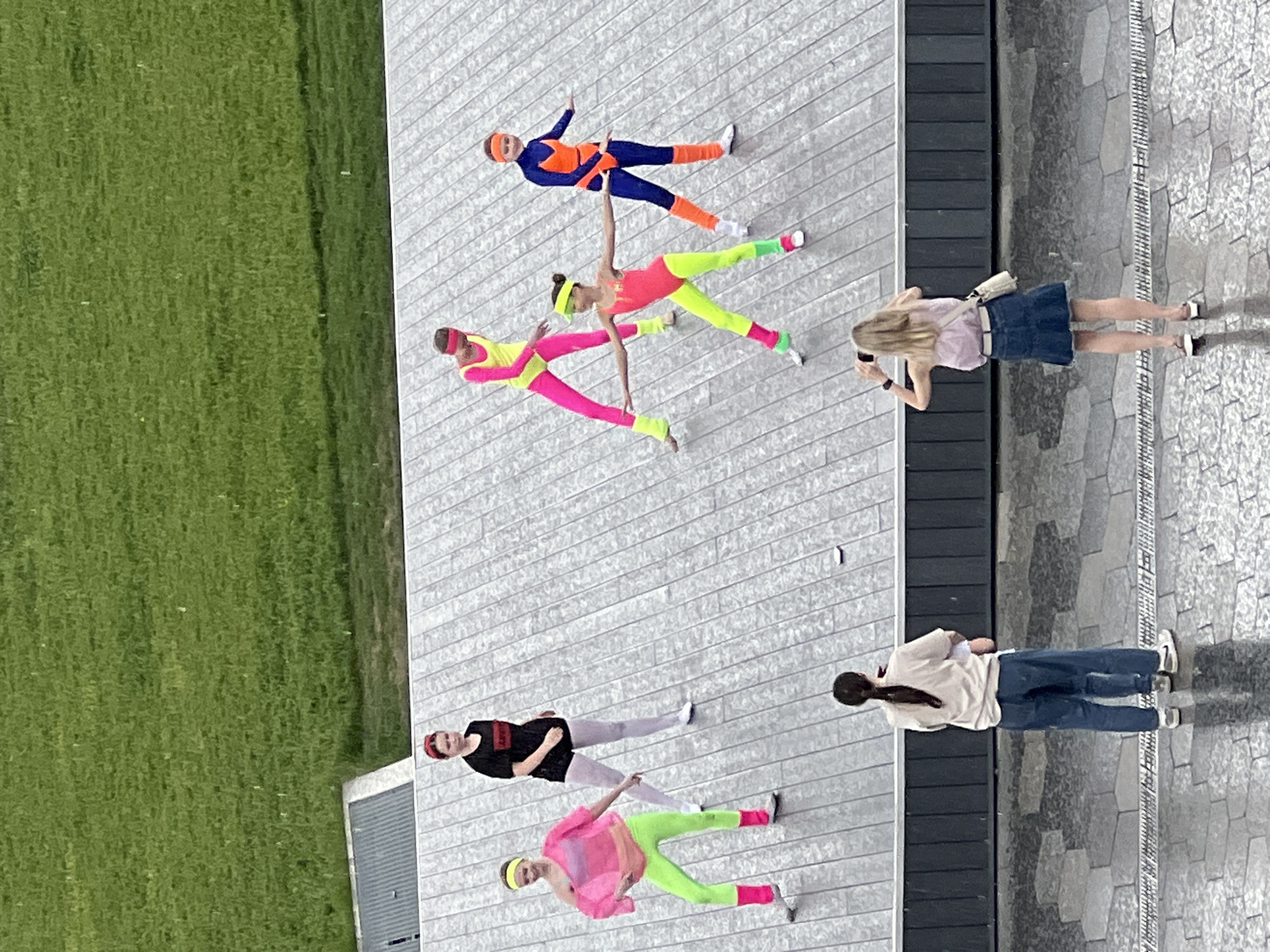 The image depicts an aerial view of a group of people on a wooden platform adjacent to a grassy area. Three people are dressed in brightly colored athletic outfits reminiscent of the 1980s, lying on the platform in poses. One person is in orange and blue, another in pink and yellow, and a third in pink and neon green. Two other individuals stand nearby, seemingly observing the scene. One is taking a photo of the athletic-clad individuals. One additional person is standing, taking notes. The location is an urban environment in Moscow, Russia, evidenced by the architectural style of the building facade to the right of the platform. The platform itself appears to be part of a public space or park area.