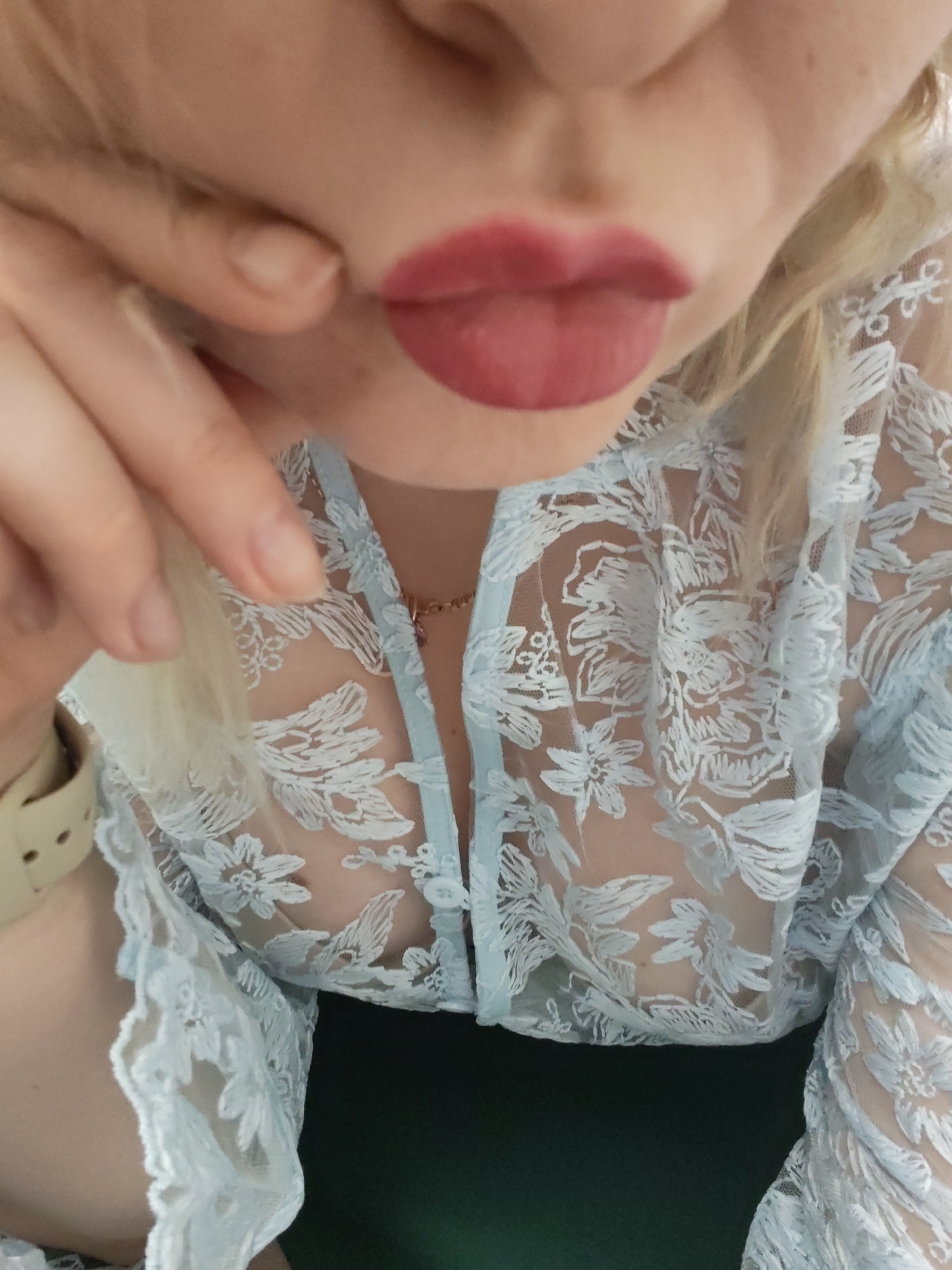 A close-up shot focuses on a person from the chest up. The subject's lips are painted with a muted red lipstick and are pursed slightly. A hand, seemingly belonging to the same person, is near their face, with a finger touching their cheek. The person is wearing a sheer, lacy top with a floral pattern. A delicate necklace is visible around their neck. A green object or fabric is seen in the lower portion of the frame, contrasting with the light colors of the top. There is no information about a specific location in Izhevsk, Russia within the frame.