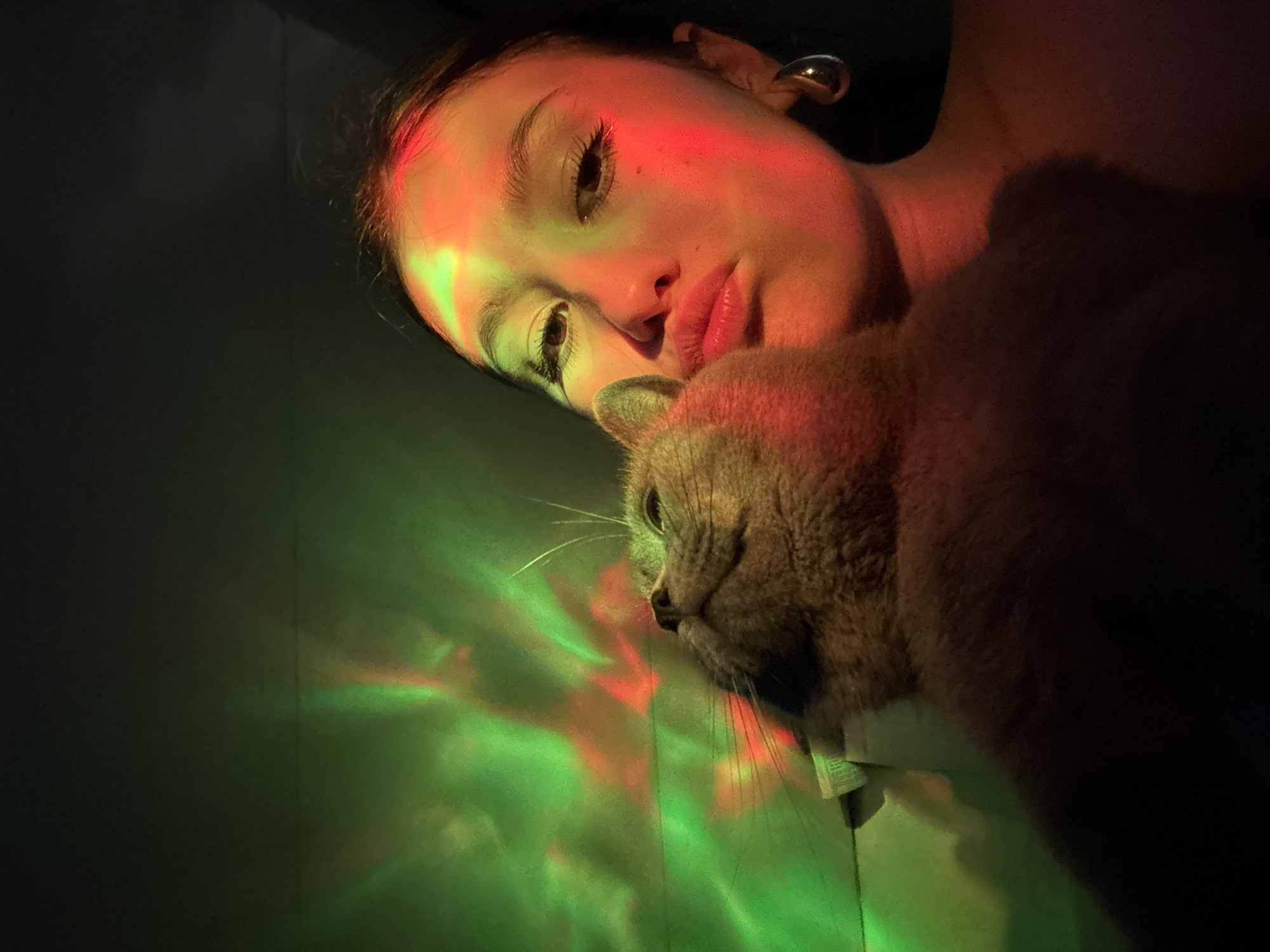 A woman lies next to a cat on a flat surface, likely a bed or floor, bathed in a mix of green and red light. The woman is light-skinned and has dark hair pulled back, with a metal earring visible. Her eyes are open, and she gazes to the left. The cat is tan-colored with green eyes and long whiskers, facing forward with its head near the woman's face. The left side of the scene is illuminated with green light, reflecting off a surface, while the right side is darker. There are no indicators to confirm the location as the city of Фоки, Russia, as there are no visible landmarks or identifying details.