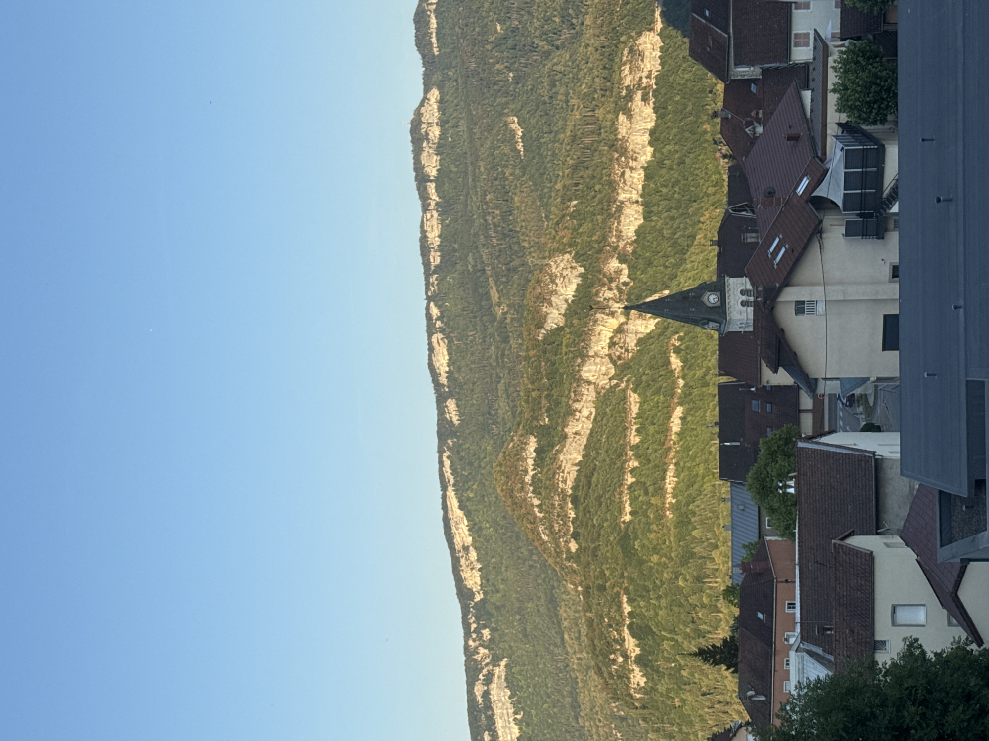The image is a vertical, medium shot of a small town in Lavans-lès-Saint-Claude, France, with a large, forested mountain range in the background. The sky is clear and blue. The town features several buildings with red tile roofs, characteristic of French architecture. A prominent church steeple with a clock is visible in the center of the town, adding a religious and historical element to the scene. The mountain range is steep and covered in dense green trees, with visible layers of rock formations. The lighting suggests it is either early morning or late afternoon, with the sun casting a warm glow on the mountain and town. There are no people visible in the image. The scene depicts a serene, rural setting, likely in the Jura region of France, known for its mountainous terrain and charming villages.