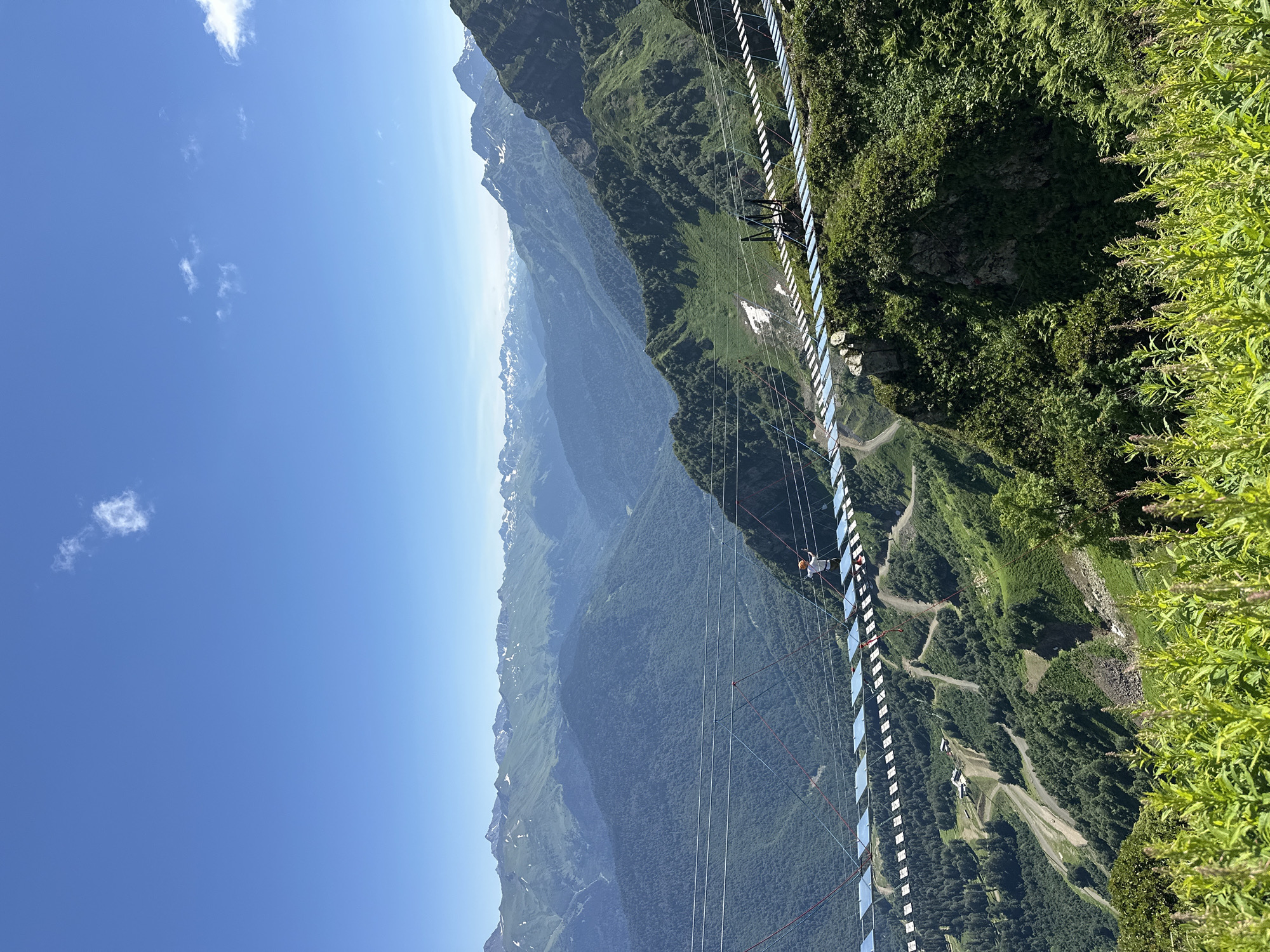 A person, wearing a white shirt and an orange helmet, is walking across a long suspension bridge, suspended between two mountains. The bridge is made of wooden planks and metal cables. The person is the only individual visible in the frame.
The scene is set in a mountainous region with lush green vegetation covering the slopes. In the background, a range of majestic mountains with snow-capped peaks stretches across the horizon under a clear blue sky. The terrain below the bridge shows winding roads and a few buildings, possibly part of a resort or ski area. The image captures a vibrant, sunny day, with a few small clouds dotting the sky.
This scene is located in Estosadok, Russia, a popular mountain resort area known for its natural beauty and outdoor recreational activities.