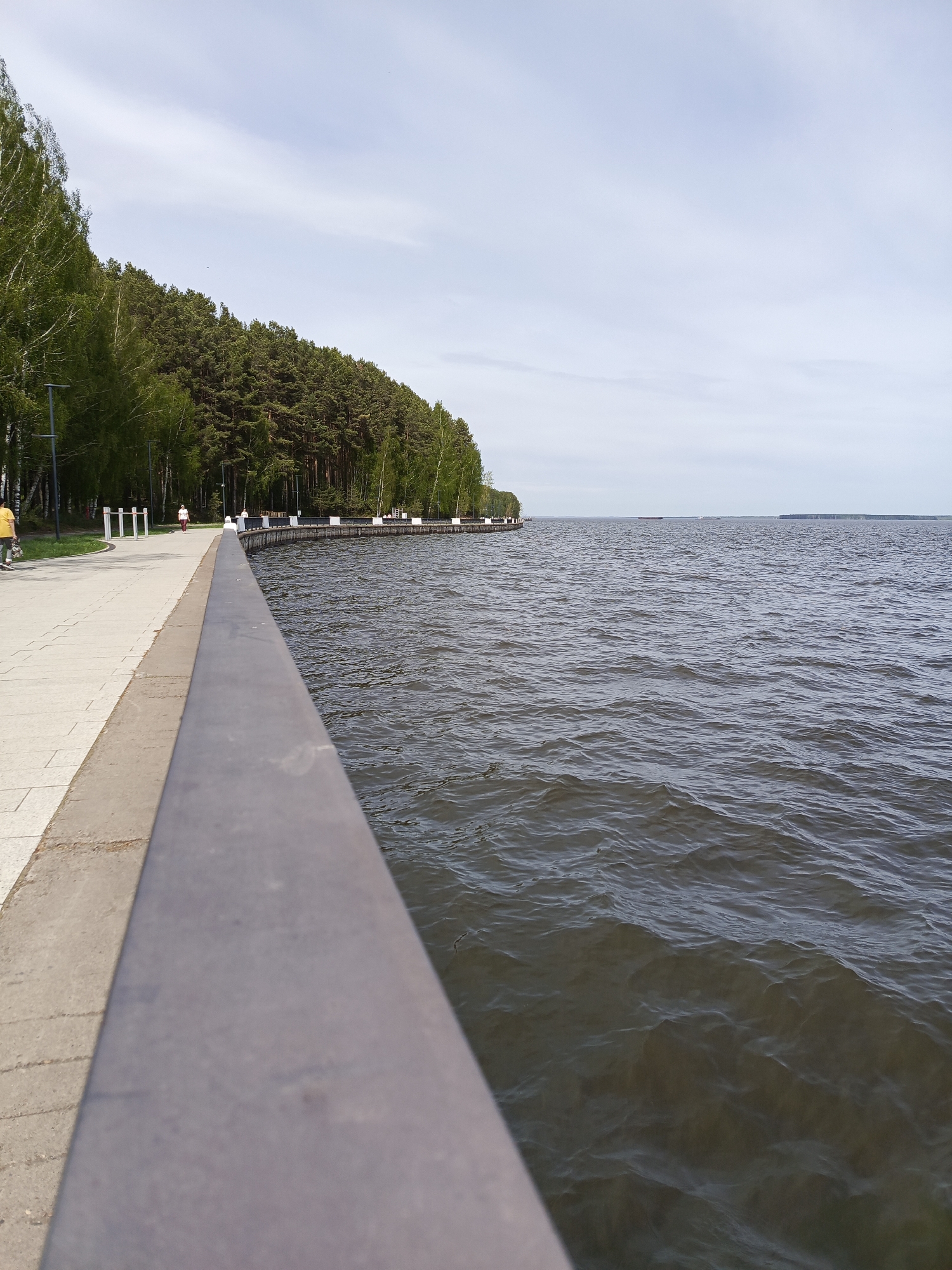 The image depicts a waterfront view from a promenade, likely in the city of Chaikovsky, Russia, based on the visual characteristics. The foreground is dominated by a gray concrete or metal railing, providing a perspective point toward the water.
To the left is a paved pedestrian walkway lined with trees and lampposts. A few people are visible in the distance along the walkway, including someone wearing a yellow jacket.
Beyond the walkway is a dense forest area along the shoreline. In the middle ground is a large expanse of water, likely a lake or a wide river. The water has a dark, slightly choppy surface with subtle ripples and waves.
In the distance, the opposite shore is visible as a low, hazy line on the horizon. The sky is overcast with a pale gray-white tone.