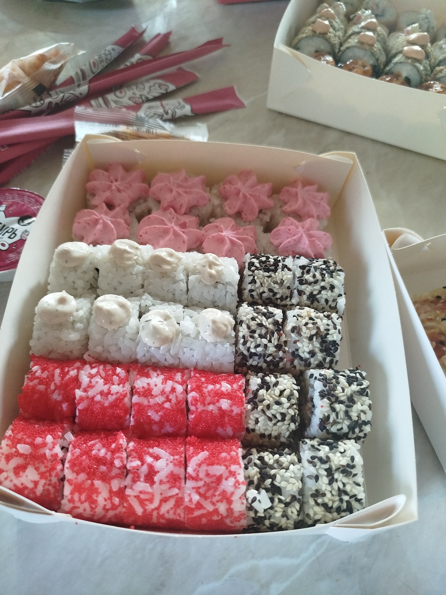 In Zavodoukovsk, Russia, a box of sushi is presented on a table, with a second box of sushi visible in the background. The primary box contains multiple rows of sushi rolls. The top row features rolls topped with pink, star-shaped dollops. The second row consists of white rice rolls with a creamy topping. The third and fourth rows display sushi rolls coated in a mixture of black and white sesame seeds. The bottom two rows showcase rolls covered in red and white tobiko (flying fish roe) or similar tiny roe. Chopsticks in pink paper wrappers are arranged on the left side of the main sushi box. A small, round container with a red lid and partially visible text is also present on the left. The setting appears to be an indoor environment, possibly a home or restaurant, with a light-colored, textured surface as the table. The lighting is ambient. No individuals are present in the frame.