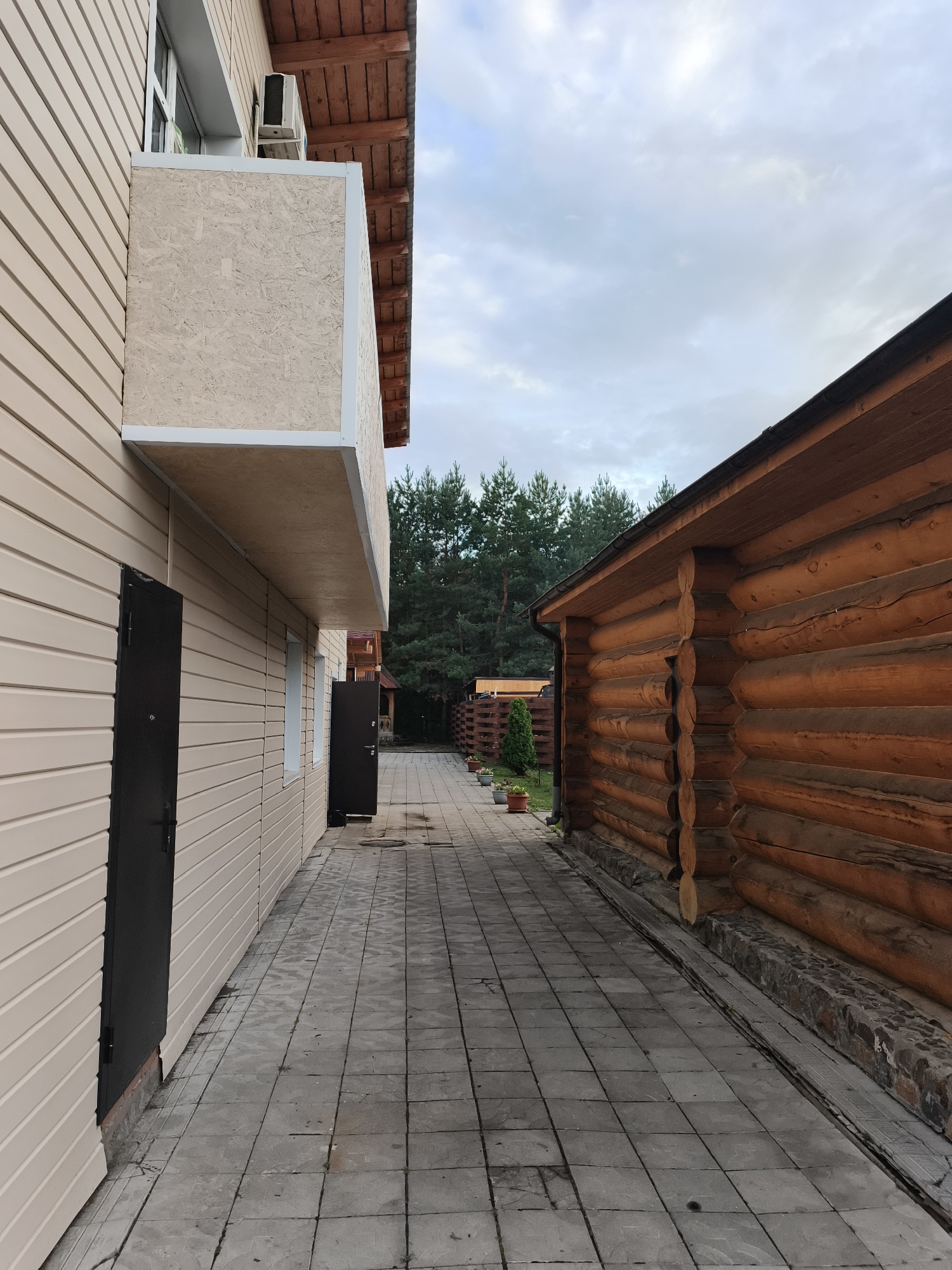 The image depicts an outdoor walkway between two buildings. On the left is a modern building with beige siding and a balcony. On the right is a traditional log cabin. The walkway is paved with square gray tiles. In the background, a dense forest of pine trees is visible under a cloudy sky. The scene appears to be set in a rural or suburban area, possibly a dacha or a resort, in the village of Katun, Russia. There are no people or animals visible in the image. The overall impression is one of a serene, natural setting.