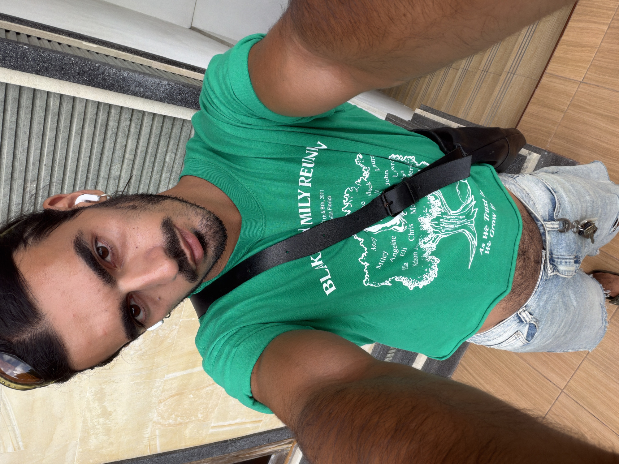 A tanned-skinned man with dark hair and a beard is captured in a selfie, wearing a green T-shirt emblazoned with "BLAK FAMILY REUNION" and a white tree design. The shirt also mentions "7th & 8th, 2011" and "ville, Florida". He's wearing a black crossbody bag with a visible strap. The man has an earbud in his left ear and is wearing denim shorts. The background consists of indoor flooring and textured grey steps. The location is identified as North Kuta, Indonesia.