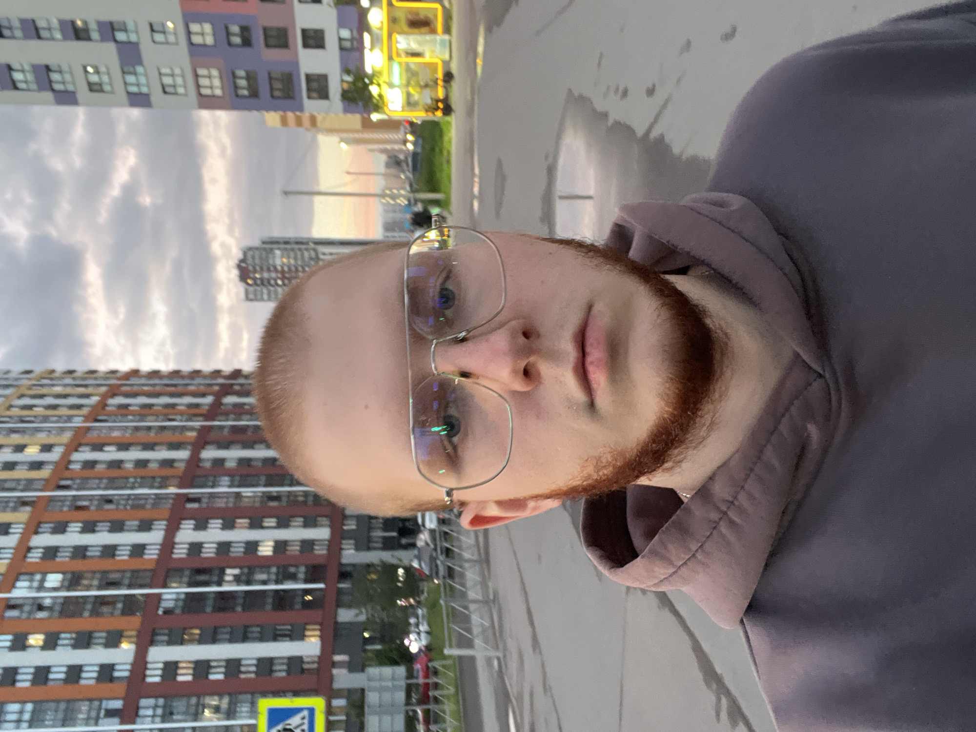 This is a close-up, selfie-style photograph of a young Caucasian male with a shaved head and a reddish beard, wearing glasses and a purple hoodie. He is standing outdoors in an urban environment. In the background, there are several tall, modern apartment buildings with colorful facades, characteristic of the residential areas in Saint Petersburg, Russia. The sky is overcast with clouds. The ground is paved and appears wet, suggesting recent rain, with puddles reflecting the surrounding buildings and sky. A yellow pedestrian crossing sign is visible to the left, and a metal fence runs along the side of the walkway. A bright yellow kiosk or food stand with illuminated signage is visible in the background on the right. The overall scene depicts a casual, everyday moment in a contemporary cityscape.
