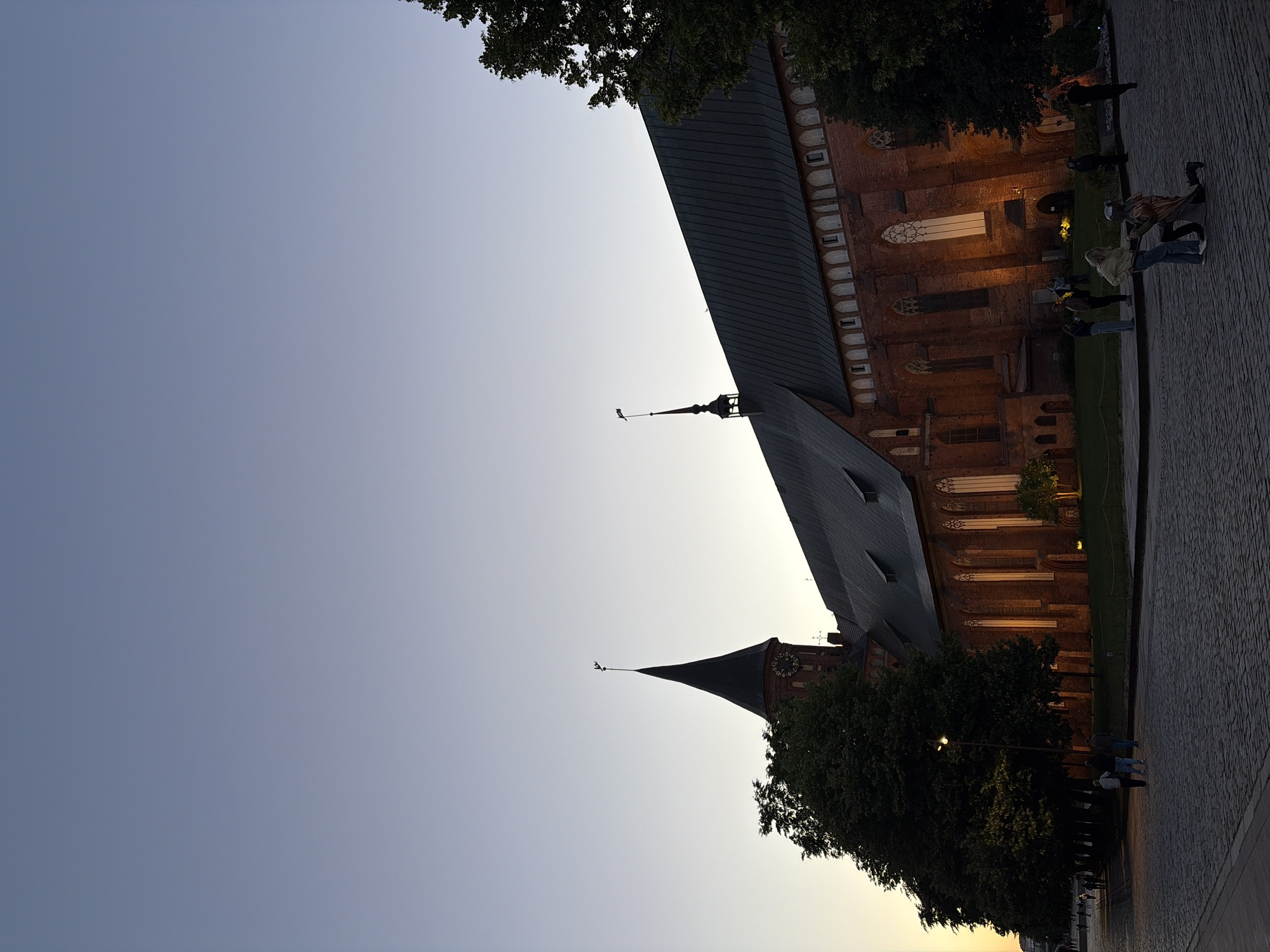 This vertical, medium-shot image shows the exterior of a large, gothic-style brick cathedral at dusk. The sky is a gradient of pale blue and gray, with the setting sun casting a warm glow on the cathedral's windows. The cathedral has a dark, sloped roof and a tall, pointed steeple with a flag on top. There is a clock on the steeple. The building is illuminated by lights, creating a warm glow. In front of the cathedral, there is a cobblestone plaza. A few trees are scattered around the plaza. Several people are visible in the plaza, some walking and others standing. One couple appears to be in the process of proposing. The scene is set in a European city.
