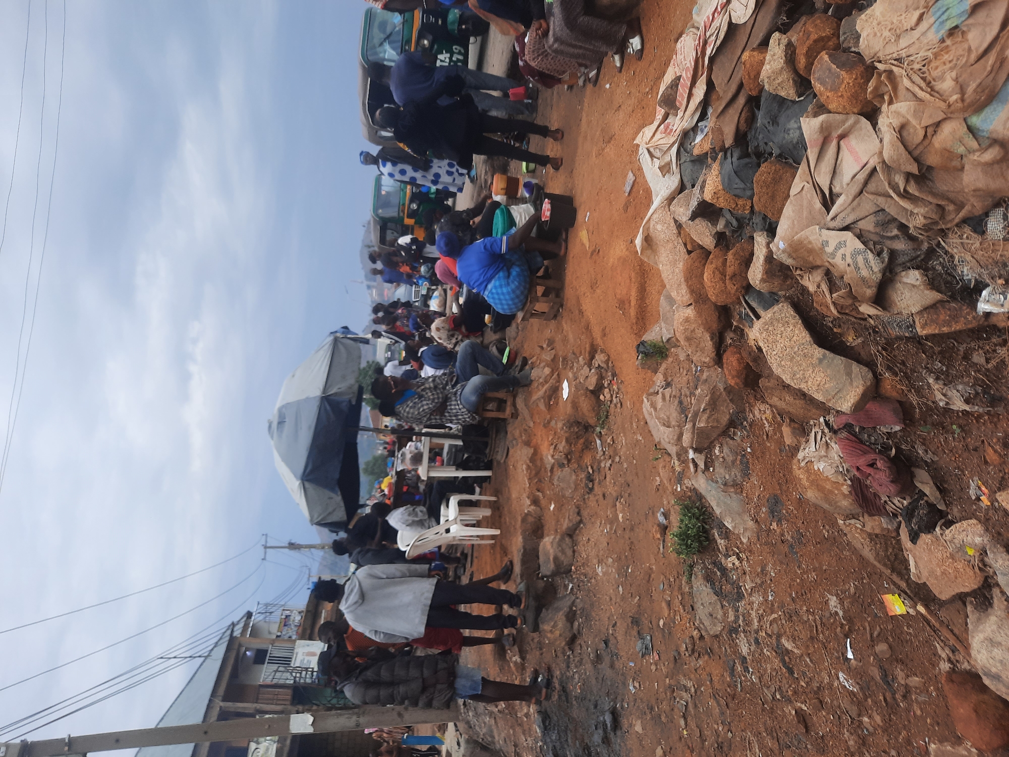 The image shows a busy roadside scene in Gwong, Nigeria. A crowd of people is gathered under a large umbrella near a row of parked vehicles, possibly taxis or buses, indicating a transportation hub. Some individuals are seated on plastic chairs, while others are standing. The ground is dirt and scattered with debris. To the right is a pile of what appears to be produce, possibly yams, covered with bags and tarps. Electrical wires are visible overhead, connecting to a nearby building. The overall scene suggests a market or gathering place in a somewhat impoverished urban environment.