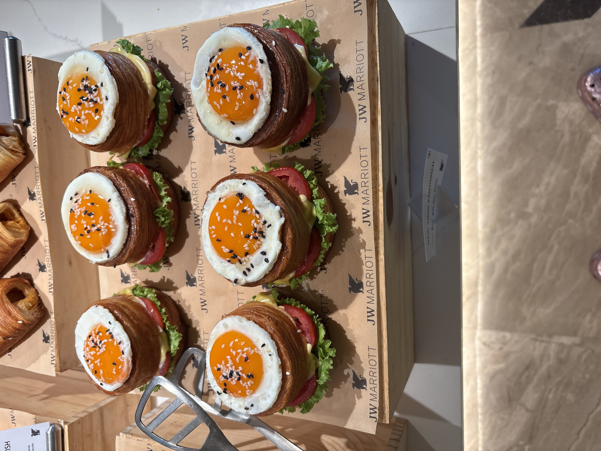 In a dimly lit display case, likely at the JW Marriott hotel in Vagator, India, a variety of pastries are presented. The foreground features six meticulously crafted "croissant burgers," each topped with a sunny-side-up egg, accented by black and white sesame seeds. Layers of sliced tomato and crisp lettuce are visible beneath the egg, nestled within a stack of flaky croissant dough. A spatulate tool rests to the left of the display. In the background, several golden-brown croissants, some appearing filled, are arranged on a textured surface bearing the "JW Marriott" logo. A small card with "NEW YORK SANDWICH" printed on it is partially visible on a lower shelf. The overall scene suggests a culinary presentation, emphasizing high-quality, artisanal baked goods.