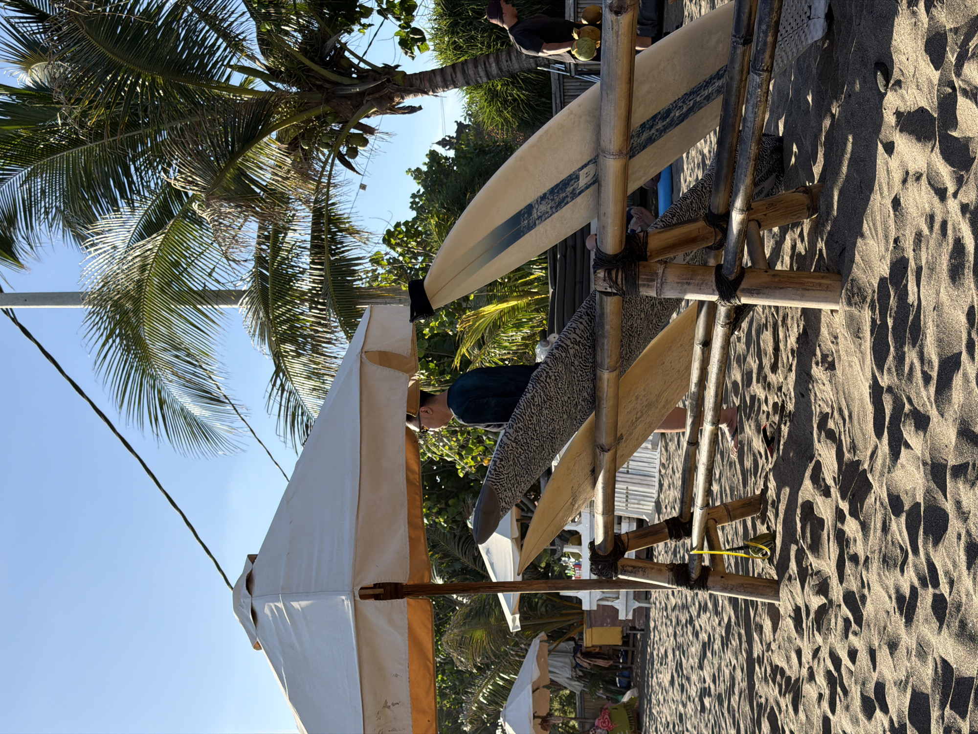 The image depicts a beach scene in North Kuta, Indonesia. There are several surfboards leaning against a bamboo structure on the sandy beach. A person with dark hair is standing near the boards. In the background, there are palm trees and other tropical vegetation. The sky is clear and blue. A white sun umbrella is also visible. The sand is light brown and textured with patterns.