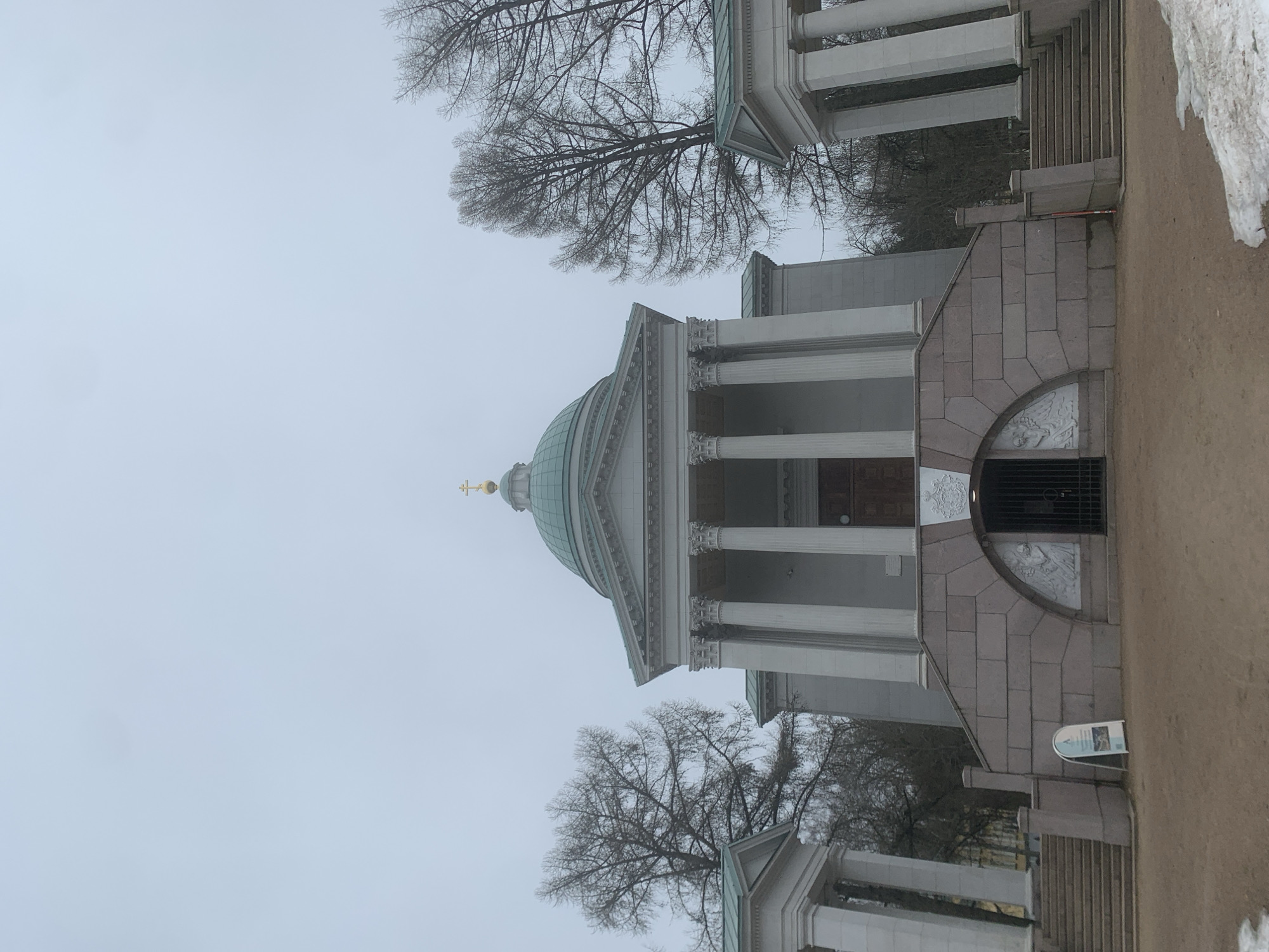 The image shows a building with a dome, columns, and a cross on top, likely a church or cathedral. The architecture suggests a classical or neoclassical style. The building has stone construction with detailed carvings and a gated entrance. There are trees with bare branches framing the building. The environment appears to be a park or outdoor area, possibly in winter or early spring due to the lack of foliage and the presence of snow. The location is Arkhangelskoye, Russia.