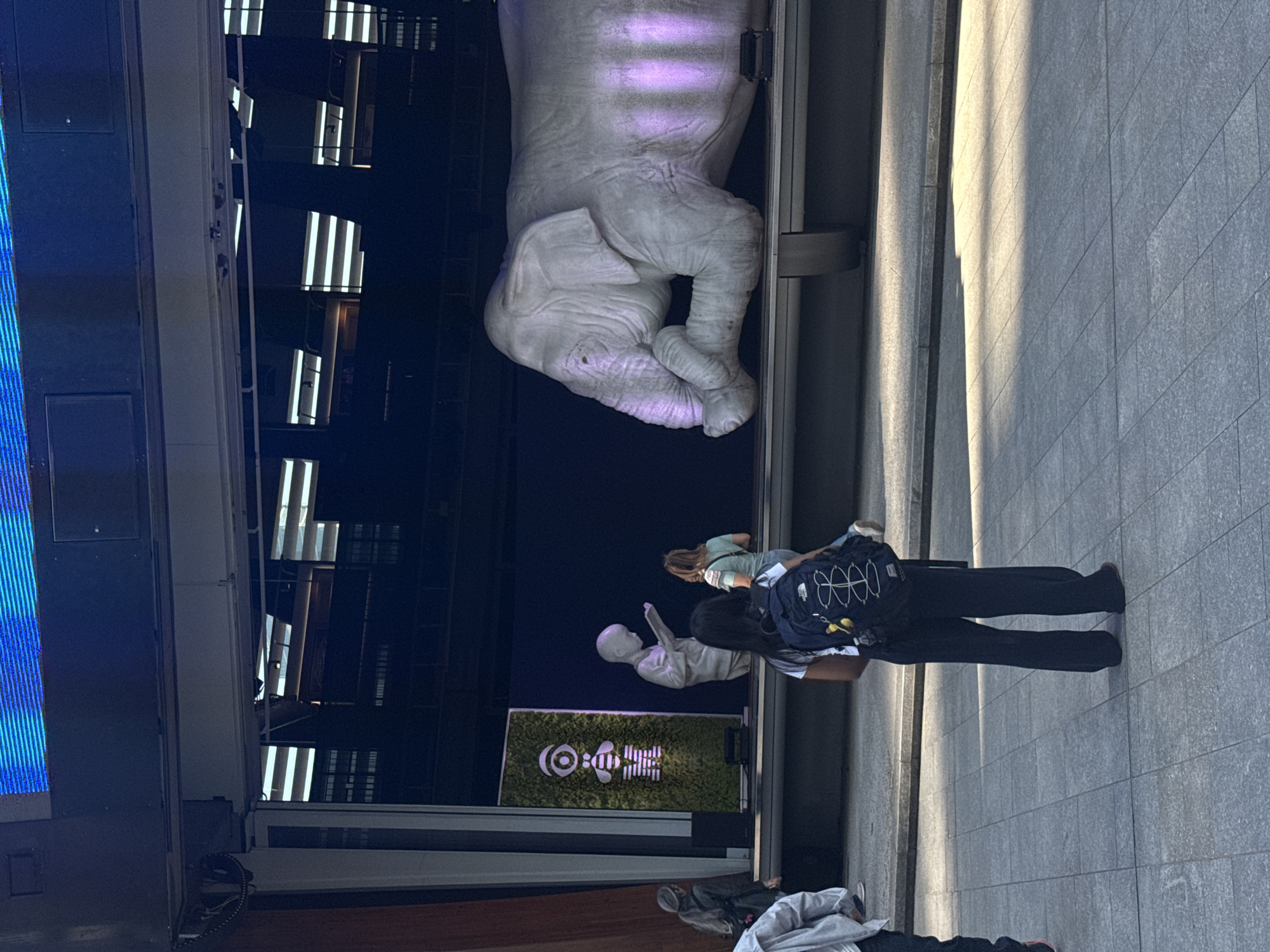 The image displays an urban scene featuring contemporary public art installations, likely in Milan, Italy. The central focus is a large, white sculpture of a human hand and forearm, emerging from a dark alcove on the left. This massive hand appears to be gently cradling a smaller, amorphous or fetal-like form, and is illuminated by a subtle purple light.
To the right of the large hand, several people are observing the art. In the foreground, a person wearing dark pants and a "THE NORTH FACE" backpack stands on a grey paved surface, facing the installations. Another individual in a light-colored top is also visible, looking towards the art. Further back, near the base of the main sculpture, is a smaller white figure, possibly a child or abstract human form, sitting on a low platform.
Behind these smaller figures, a prominent vertical screen or sign with a bright green background displays white graphics. These graphics include a circular logo featuring a stylized bee-like creature and some stylized, partially visible text.
The background on the far left shows reflections of vertical strip lights from what appears to be a building's interior or a digital display. The lighting on the paved ground to the right suggests it is daytime or a very well-lit covered area. The overall atmosphere is one of casual observation and engagement with modern art in a public space.